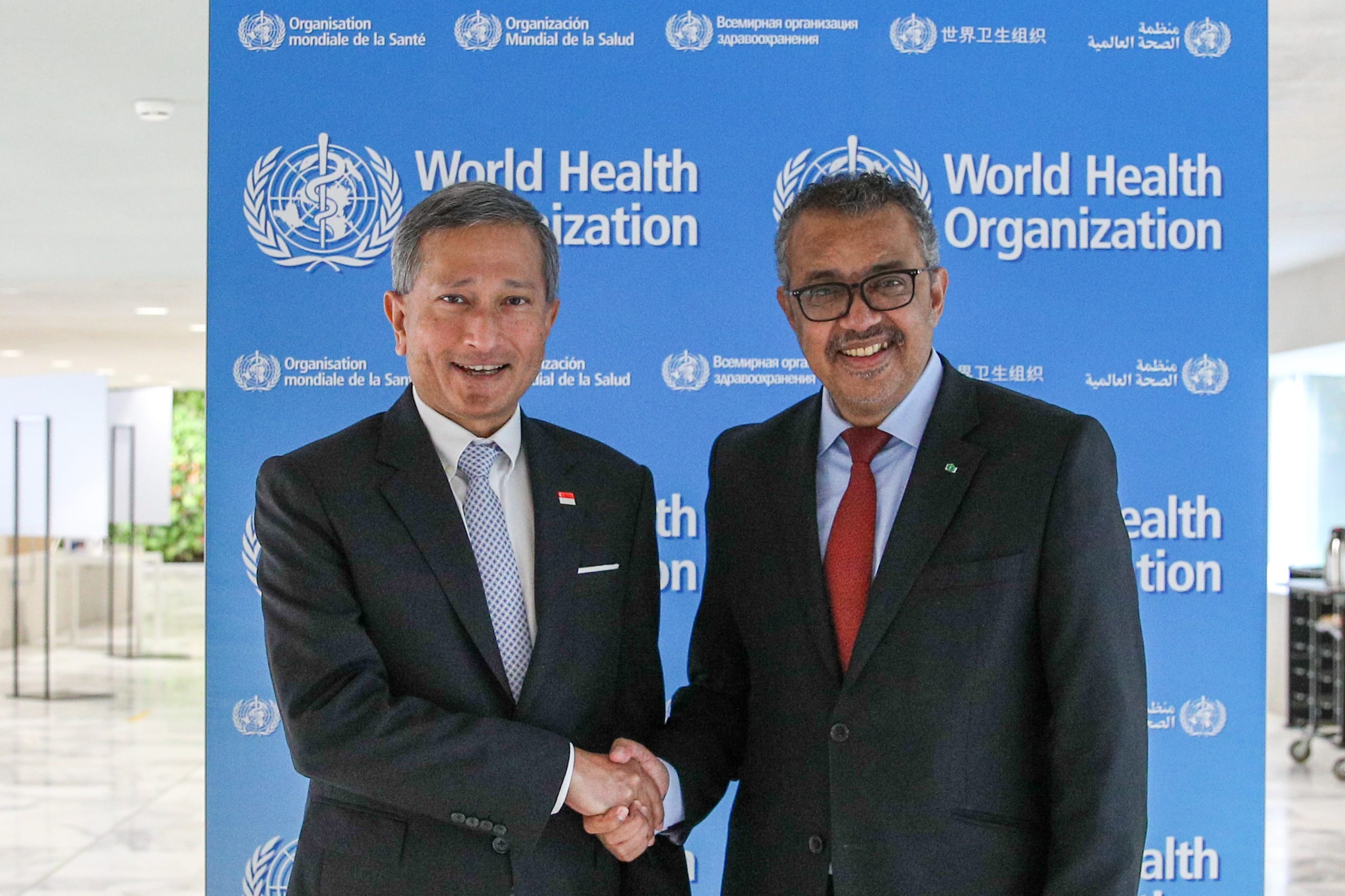 Two men in suits shake hands in front of a blue WHO banner.