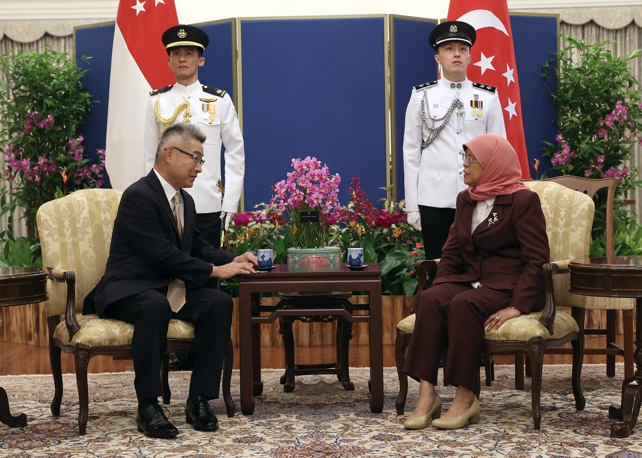 Two people seated with guards and Singapore flags in the background.
