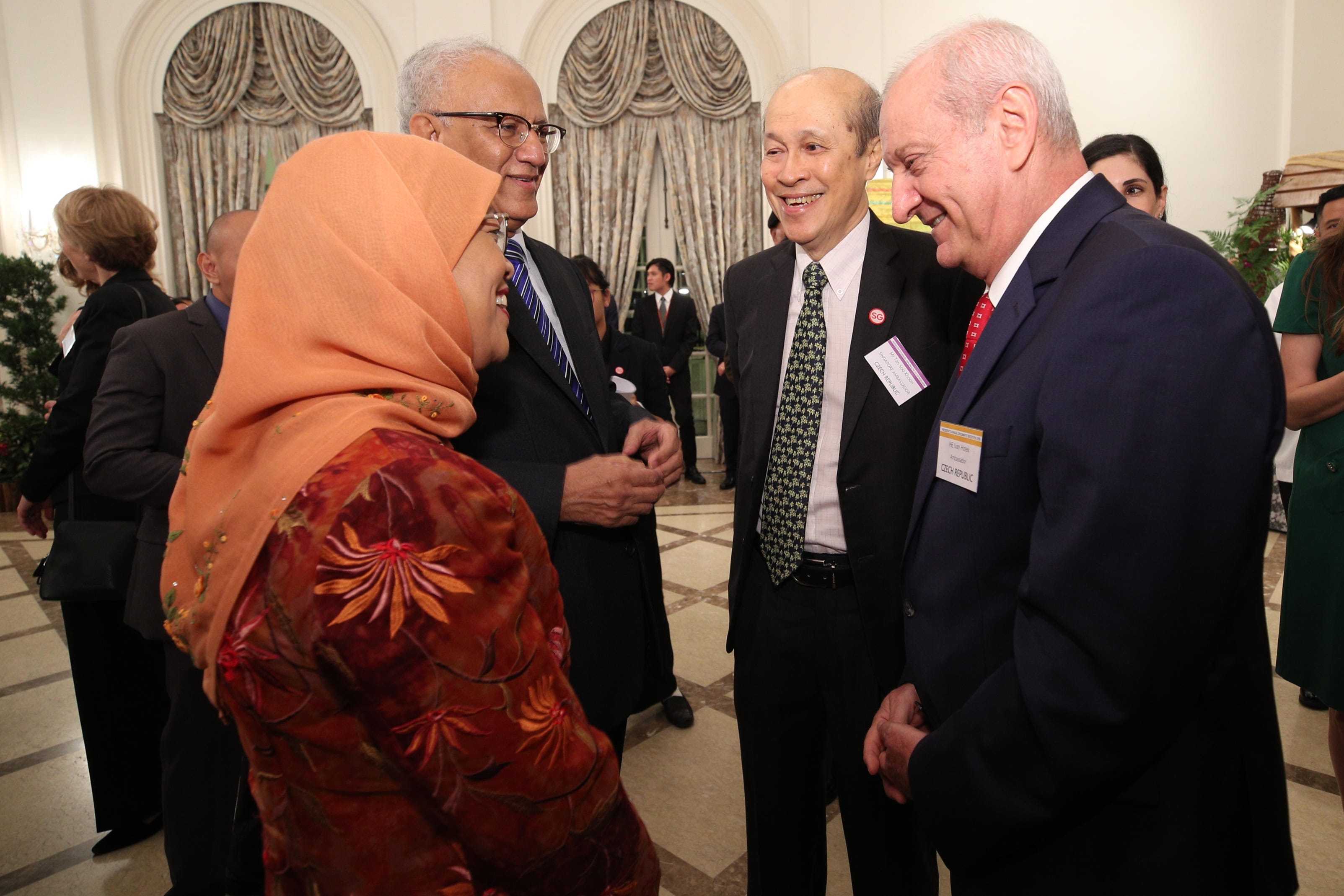 Group of people in suits and a woman in hijab indoors, talking. Nametags visible.