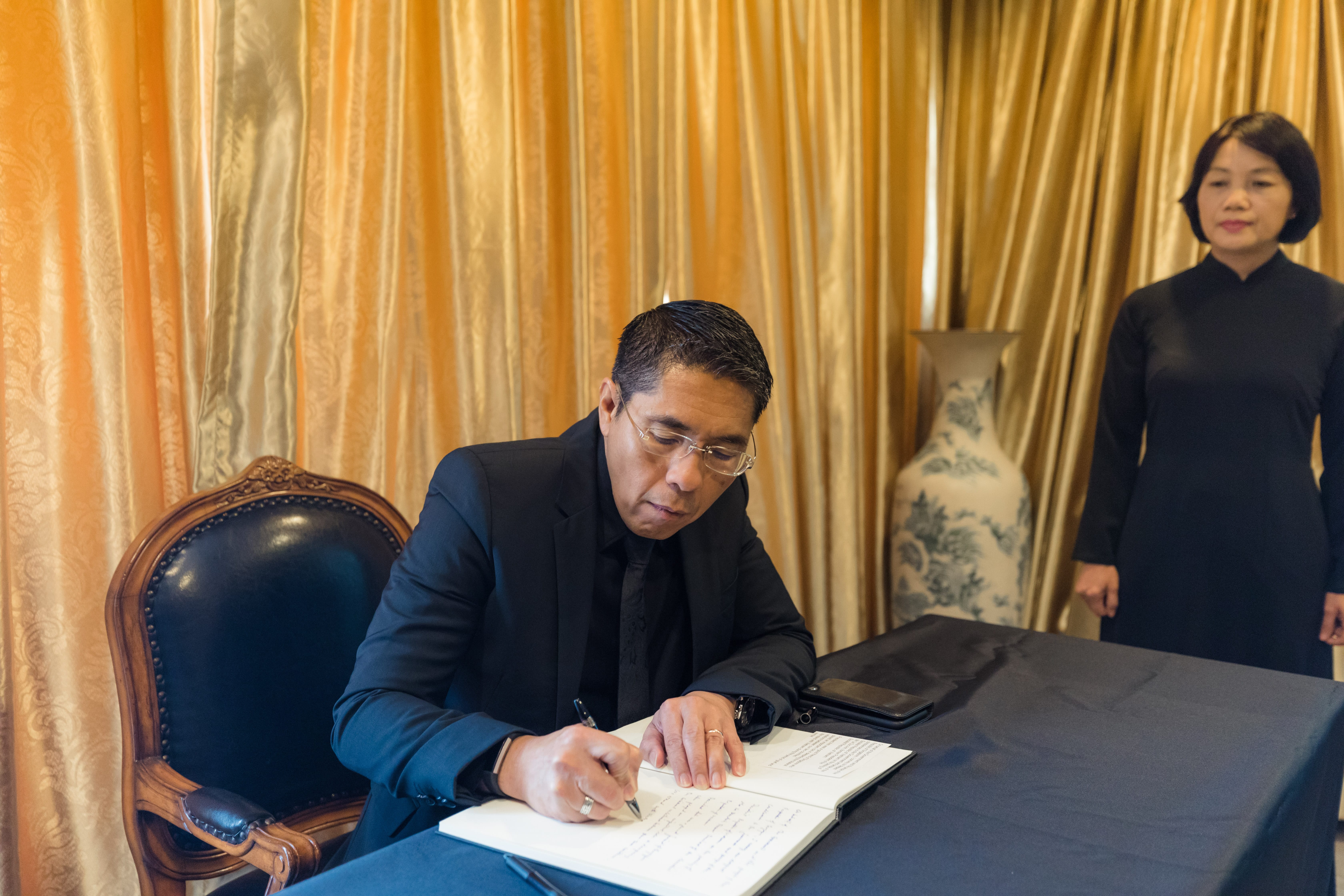 Person signing book at table, another person in background. Gold curtain backdrop.