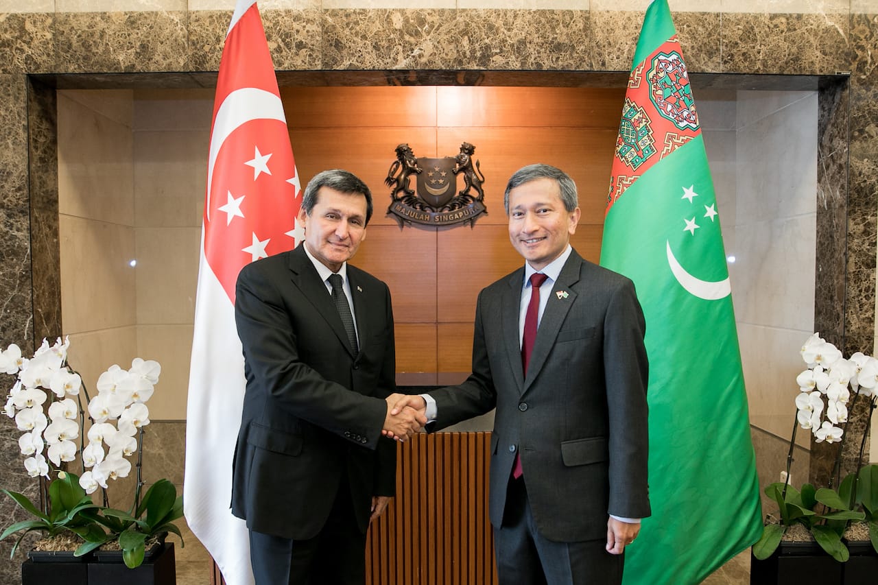 Two men in suits shake hands between the Singapore and Turkmenistan flags.