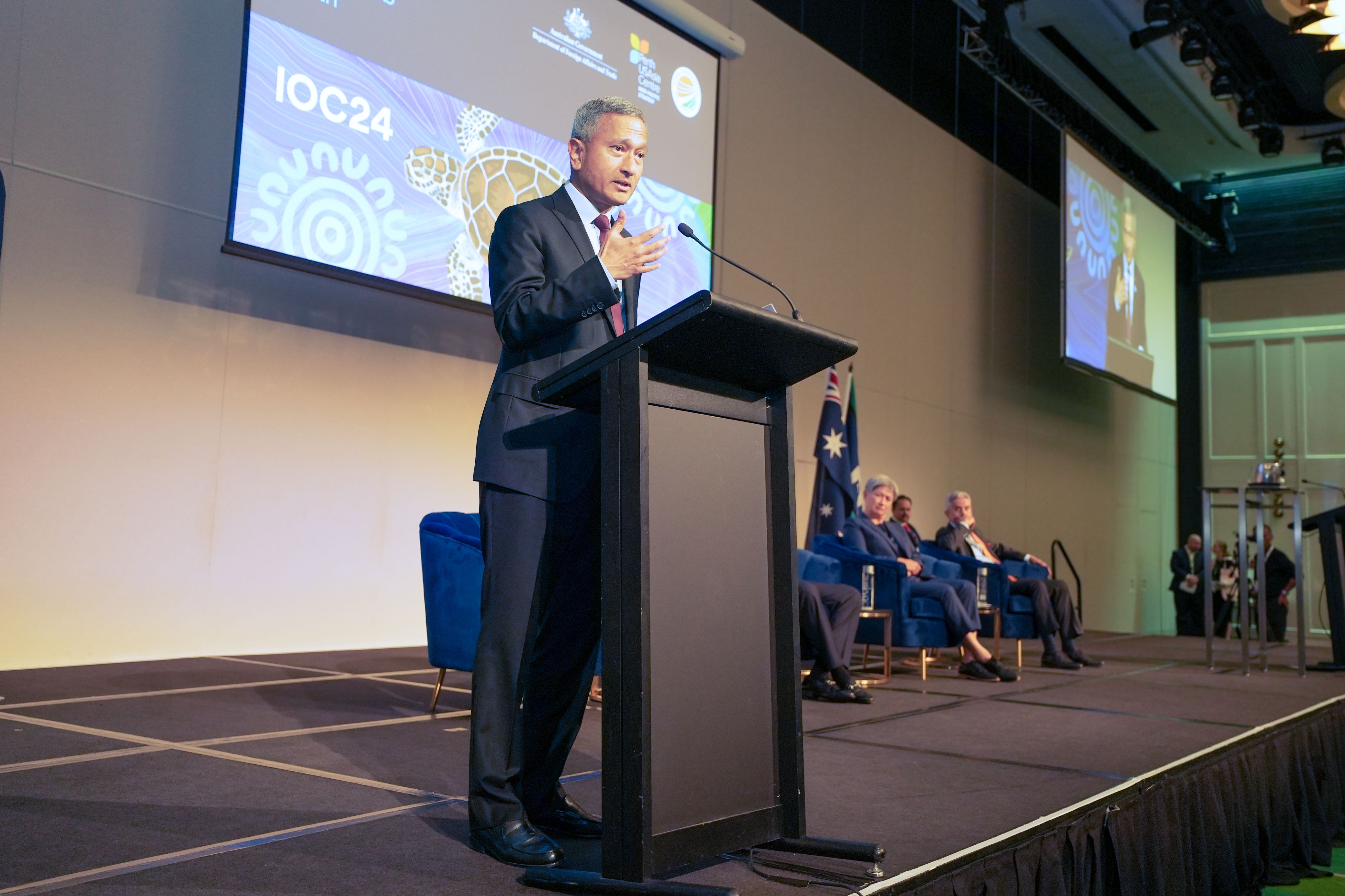 Speaker at podium with IOC24 logo, Australian flag behind seated panelists.