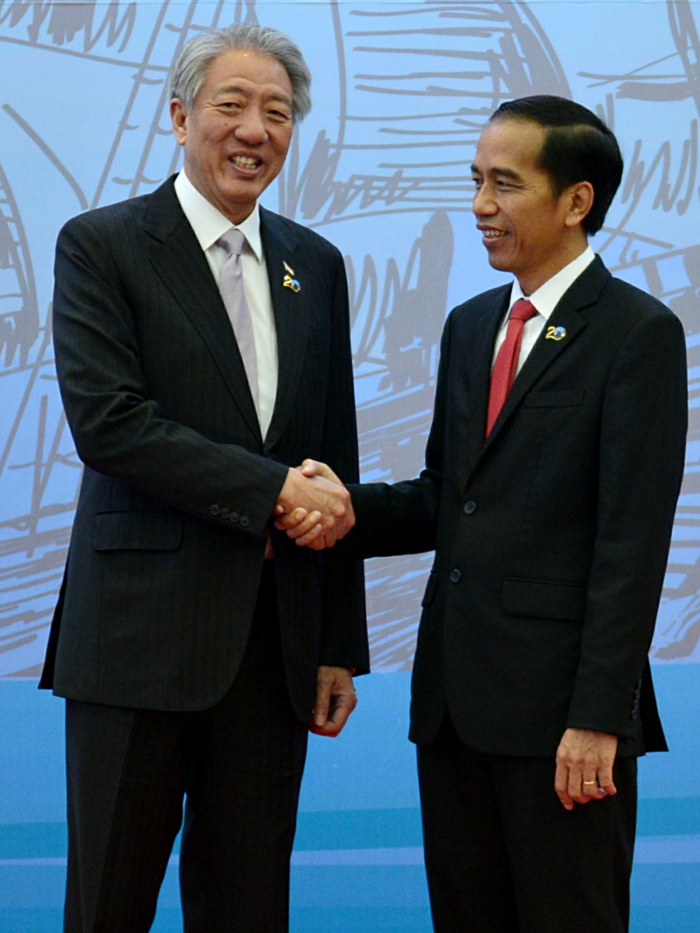 Two men in suits, Jokowi on the right, shake hands in front of a blue background.