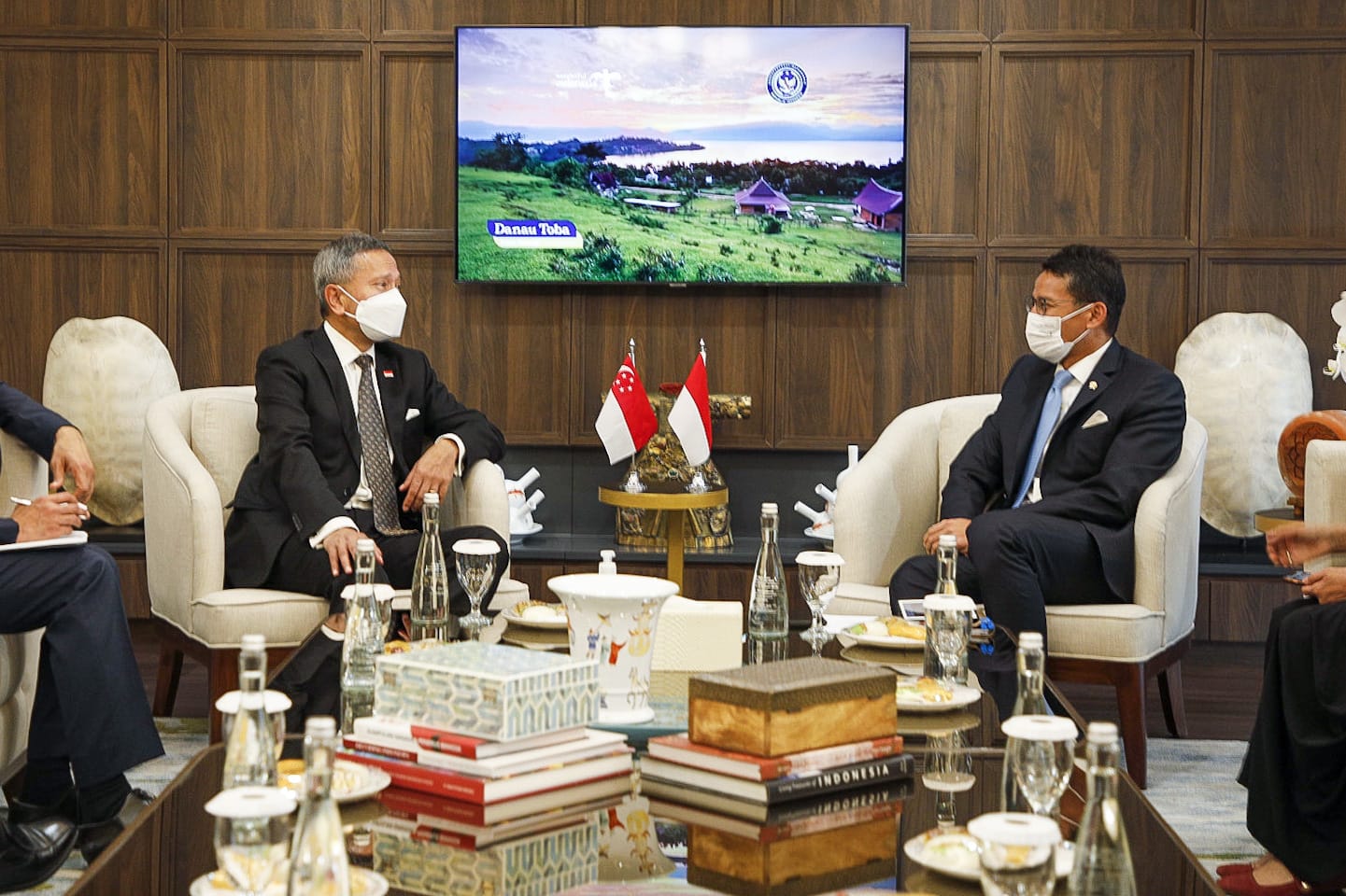 Two men in suits wearing masks sit facing each other near Singapore & Indonesia flags. TV shows Danau Toba scenery.