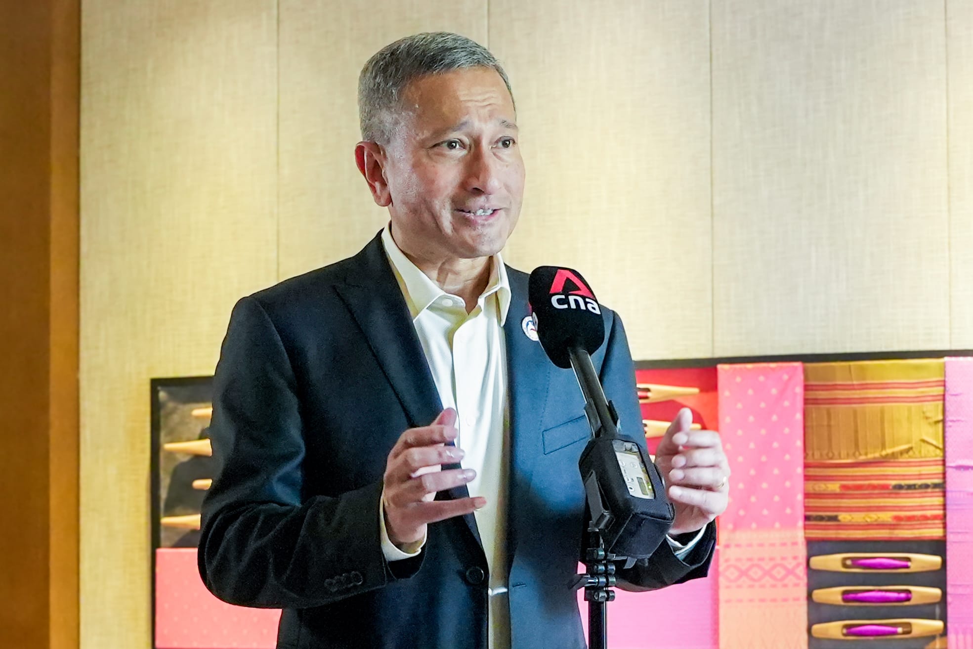 Man in suit speaks into CNA microphone, hands gesturing. Decorative fabric art behind.
