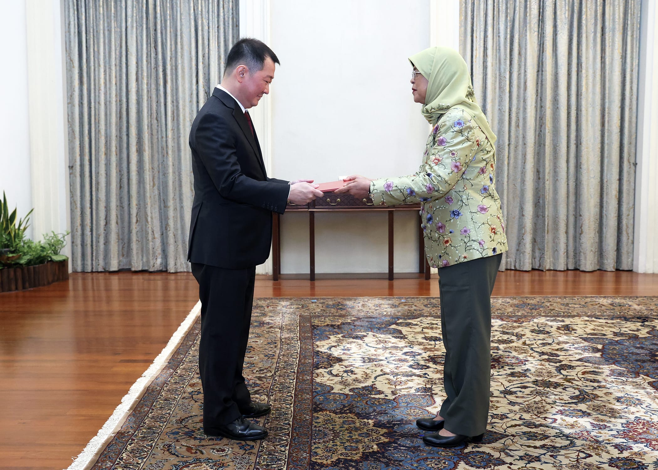 Two people exchanging a red document, indoors on an ornate rug near patterned curtains.