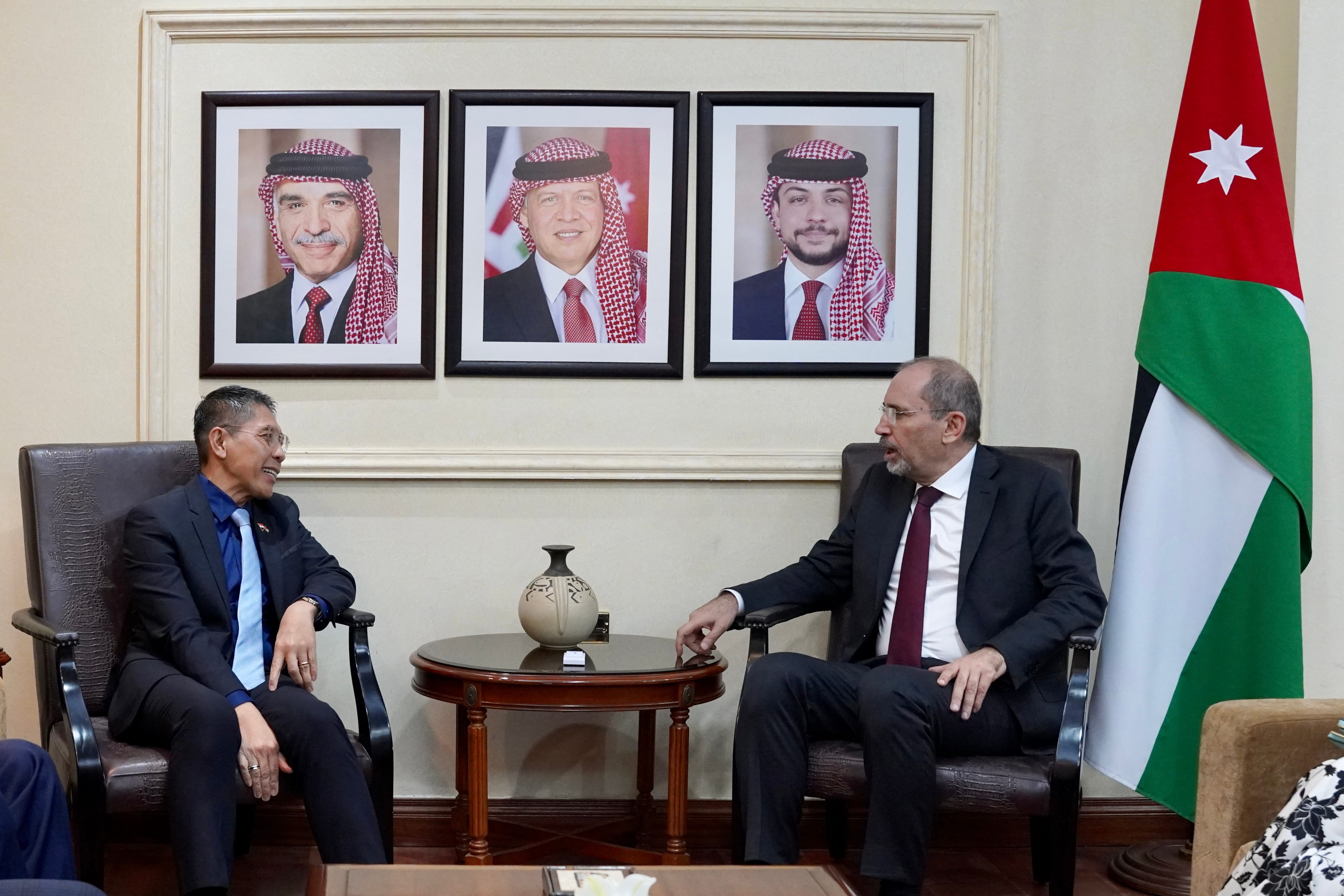 Two men in suits seated with Jordanian flag and framed portraits of royalty in background.