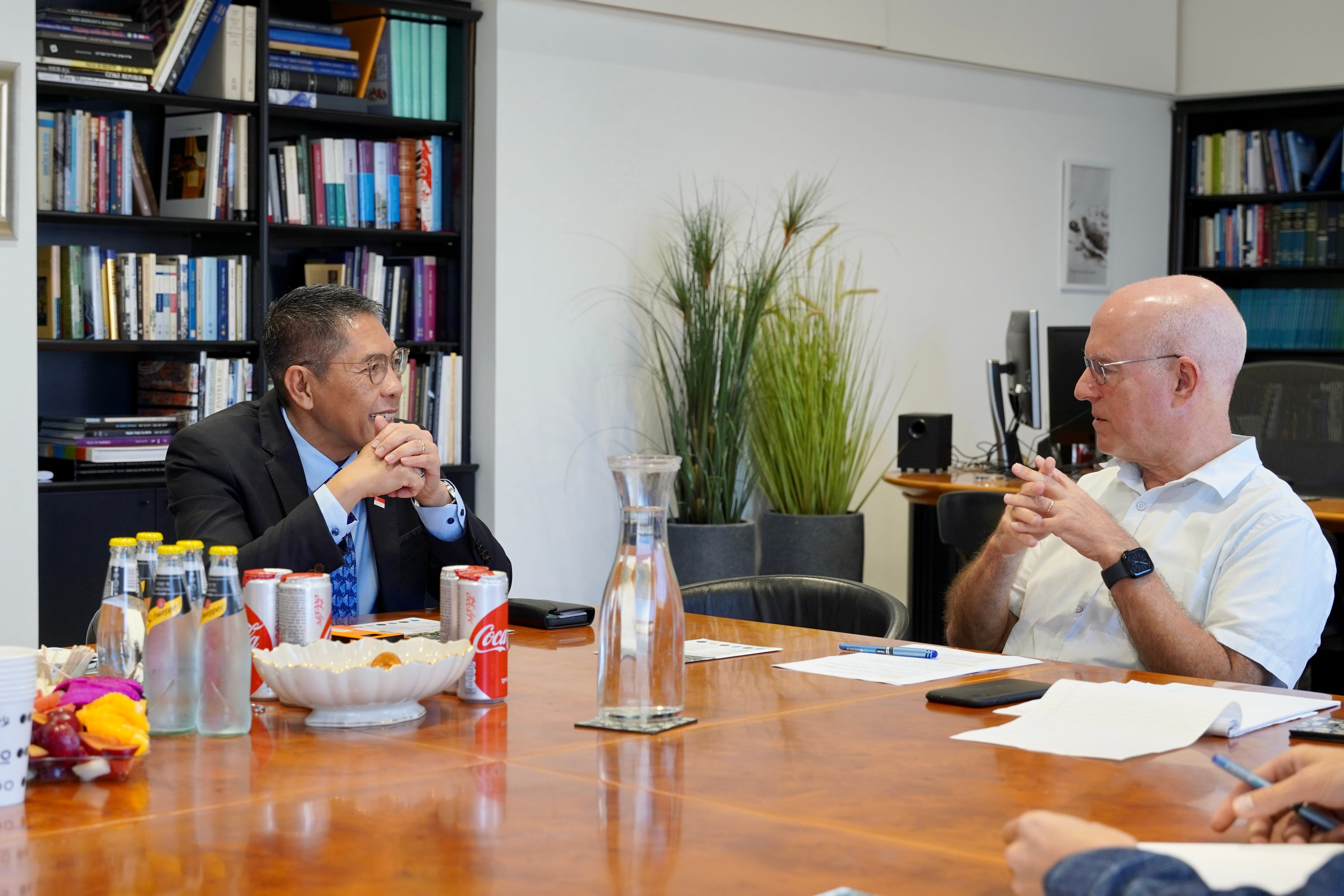 Two men sit at a table with drinks, a bowl, and documents. Bookcases visible.