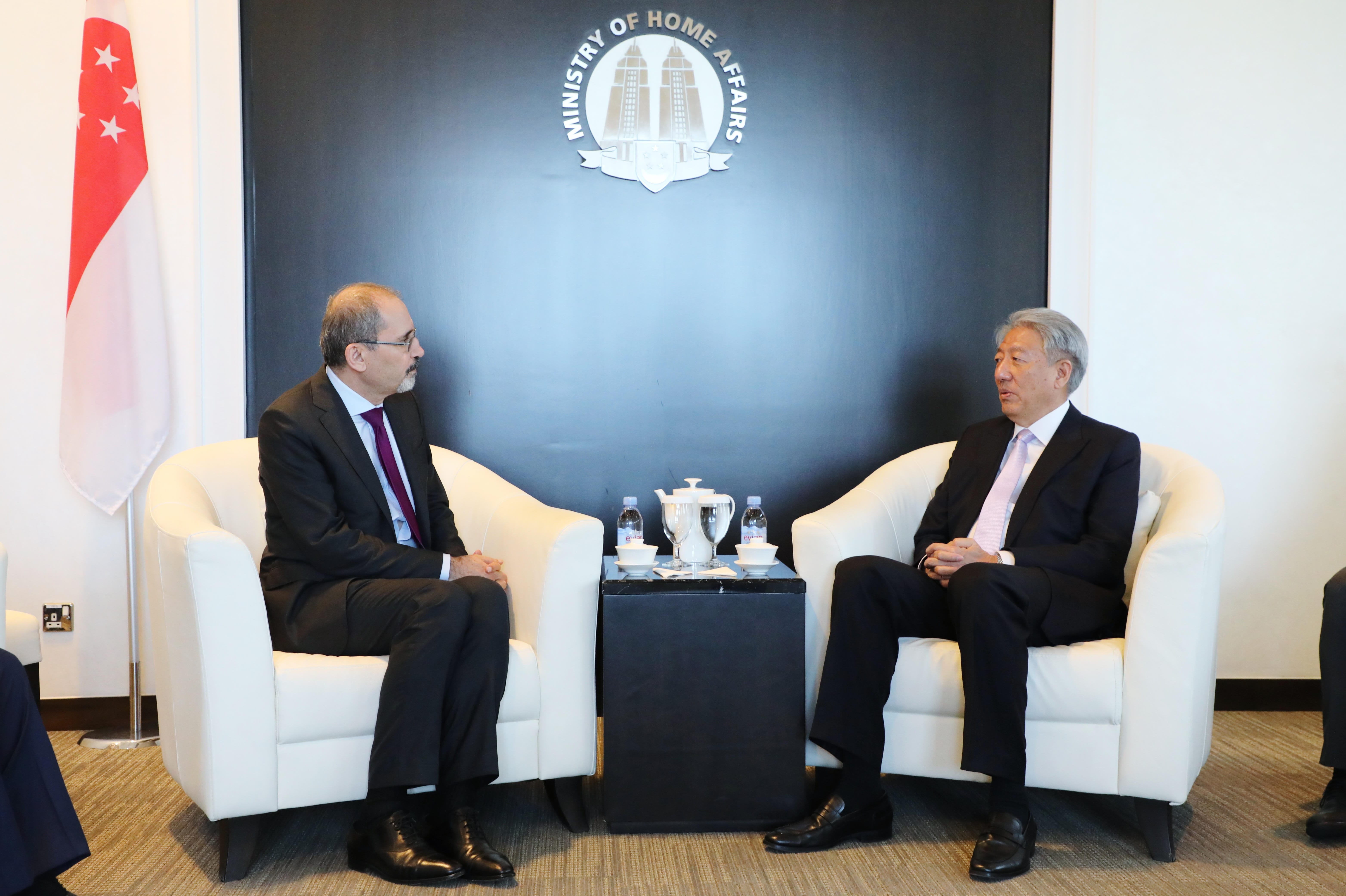 Two men hold a gift-wrapped box in a room with framed portraits and Kuwaiti flag.