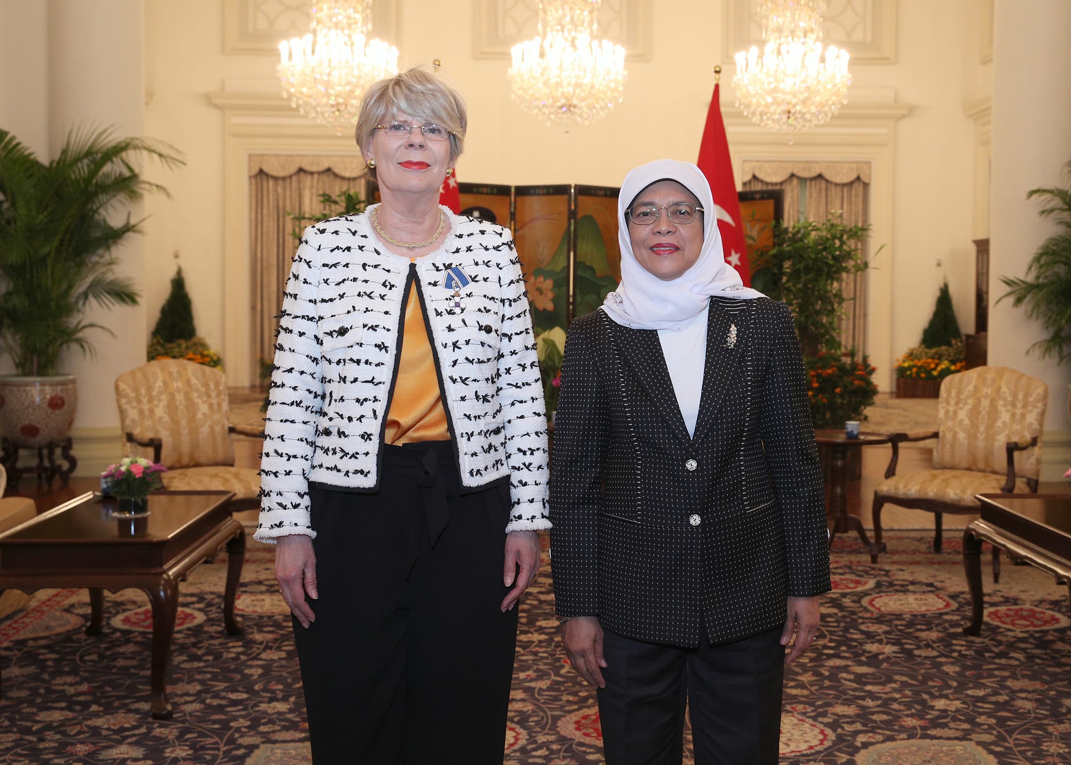 Two women stand in a room with chandeliers and a Turkish flag in the background.