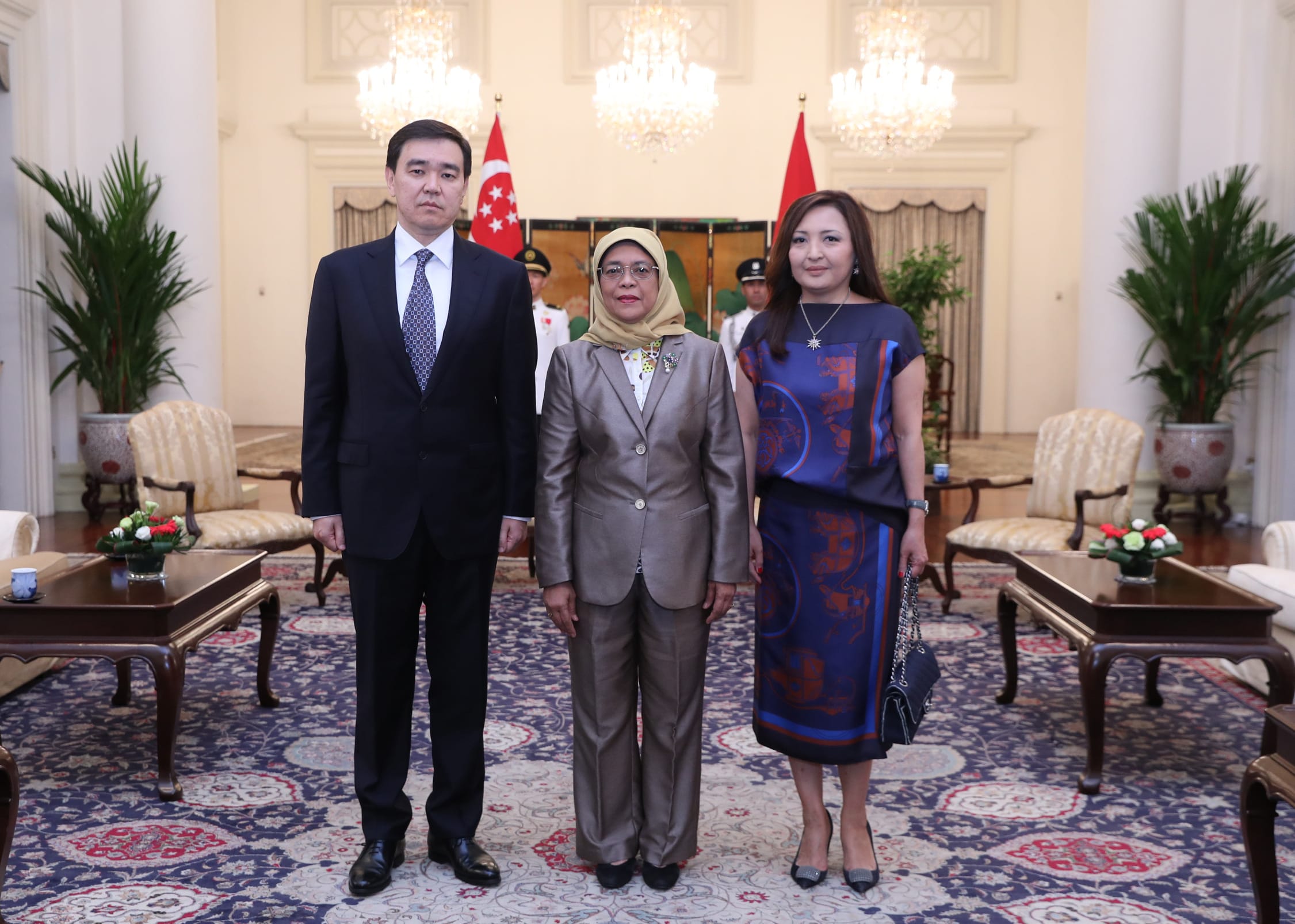 Three people in formal attire stand in a room with Singaporean flags.