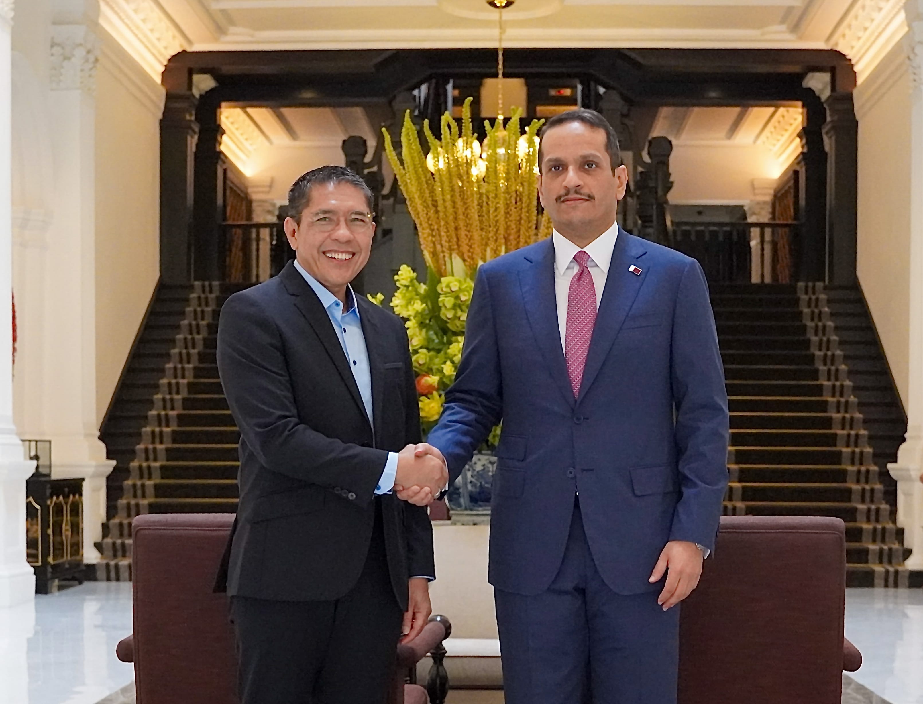 Two men in suits shaking hands in a grand hall, staircase visible behind them.