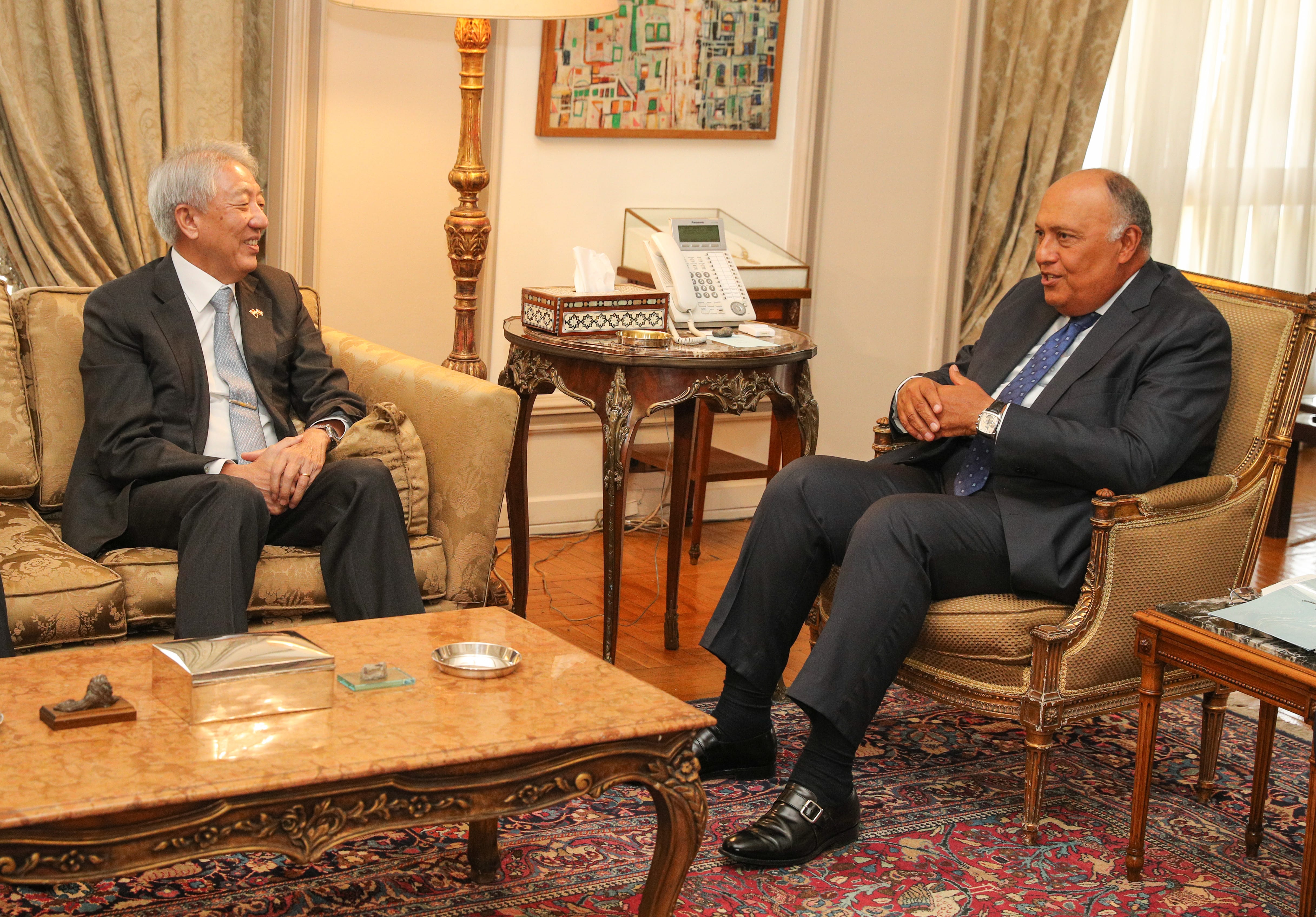Two men in suits seated in ornate room, engaged in conversation.