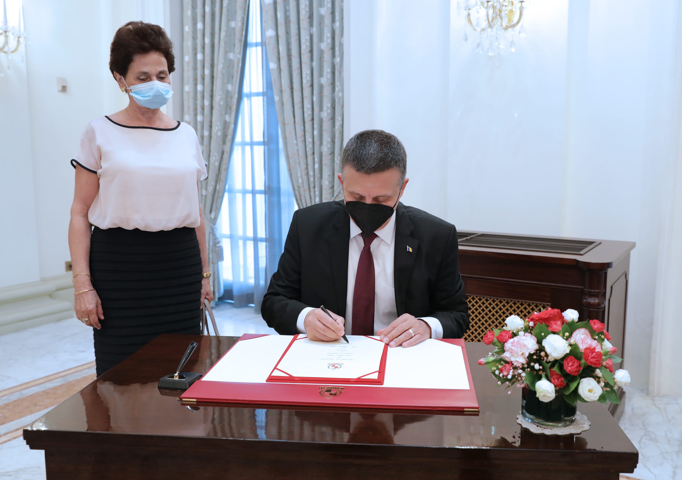 Man in suit and face mask signing document; woman in mask stands nearby.