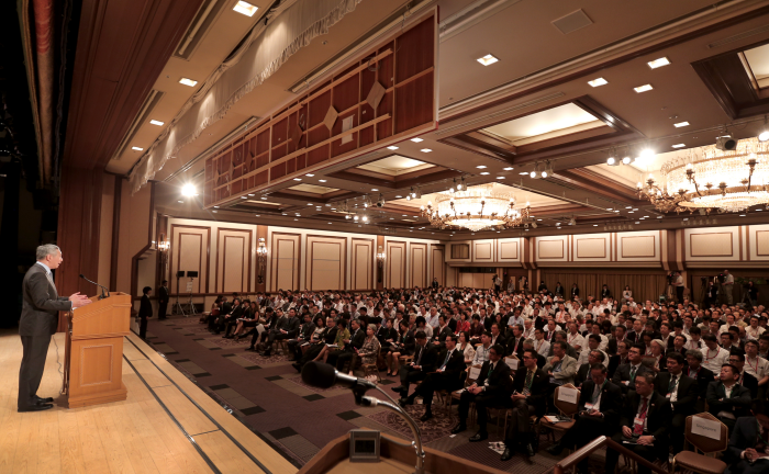 Man speaking at podium to a large seated audience in a grand ballroom.