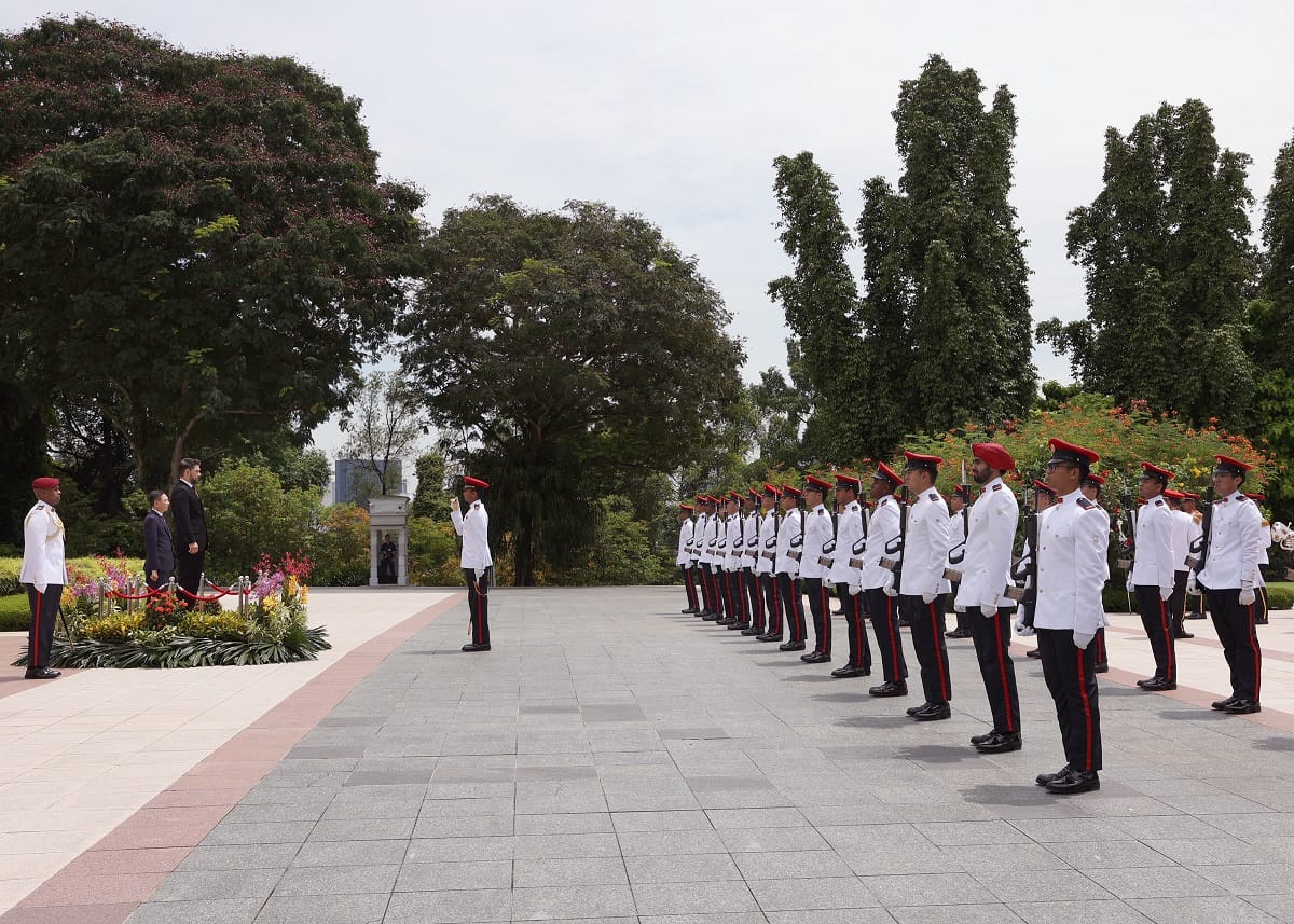 Formal outdoor scene with men in military uniform and suits standing in rows on tiled plaza.