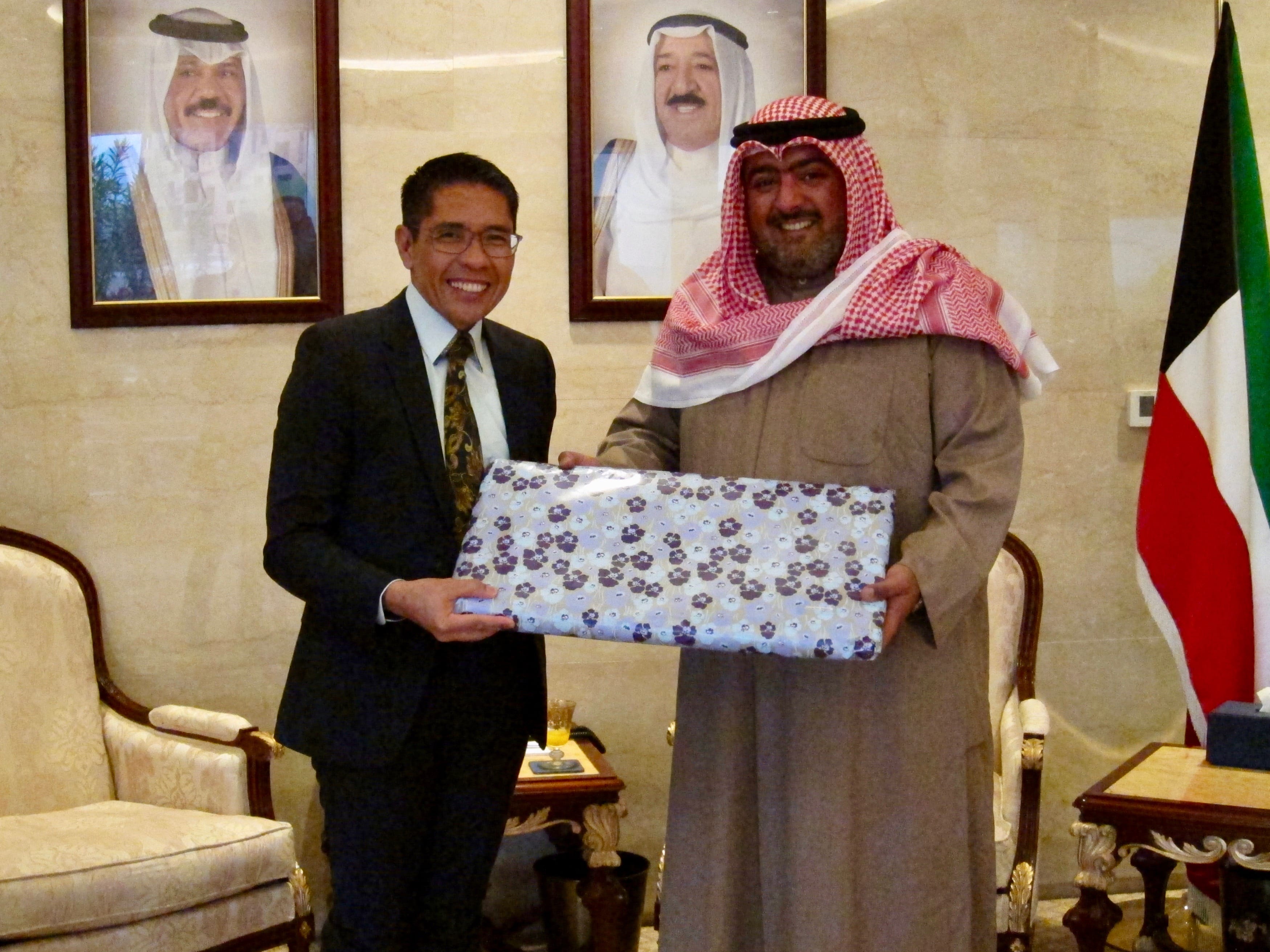 Two men holding a gift wrapped with floral paper, portraits and Kuwait flag behind them.