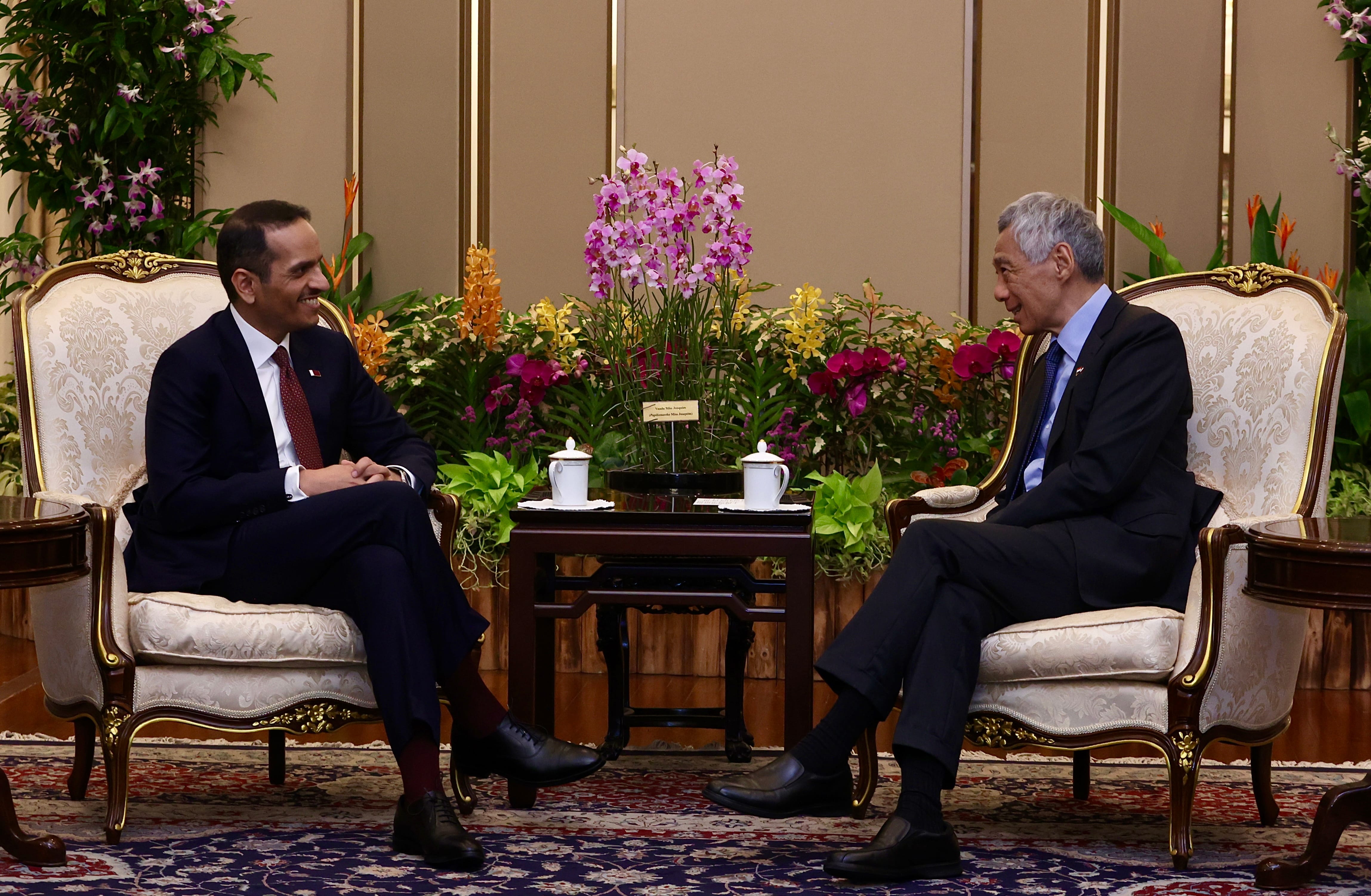 Two men in suits sit in ornate chairs facing each other, flowers between them.