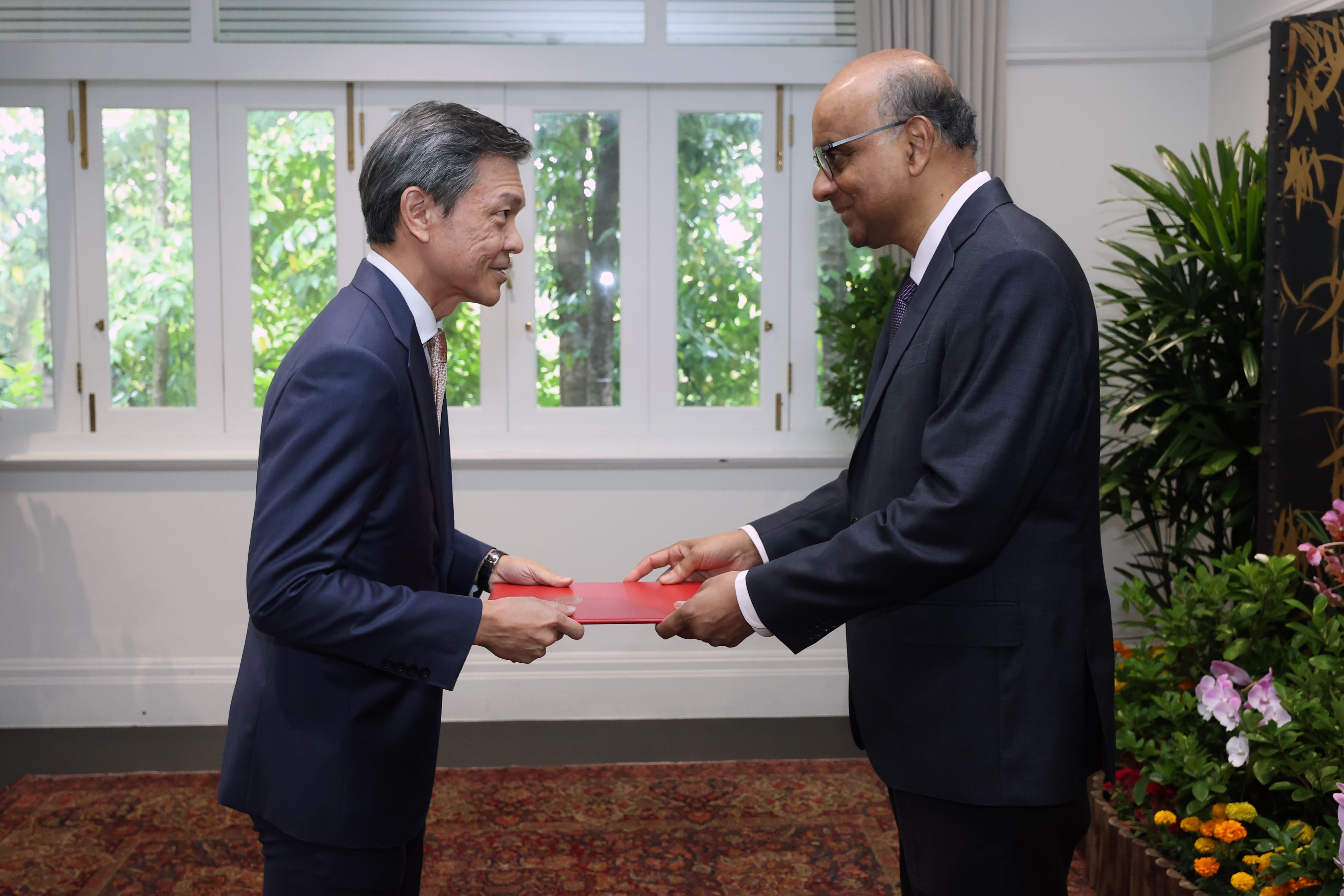 Two men in suits exchanging a red folder indoors near a window.