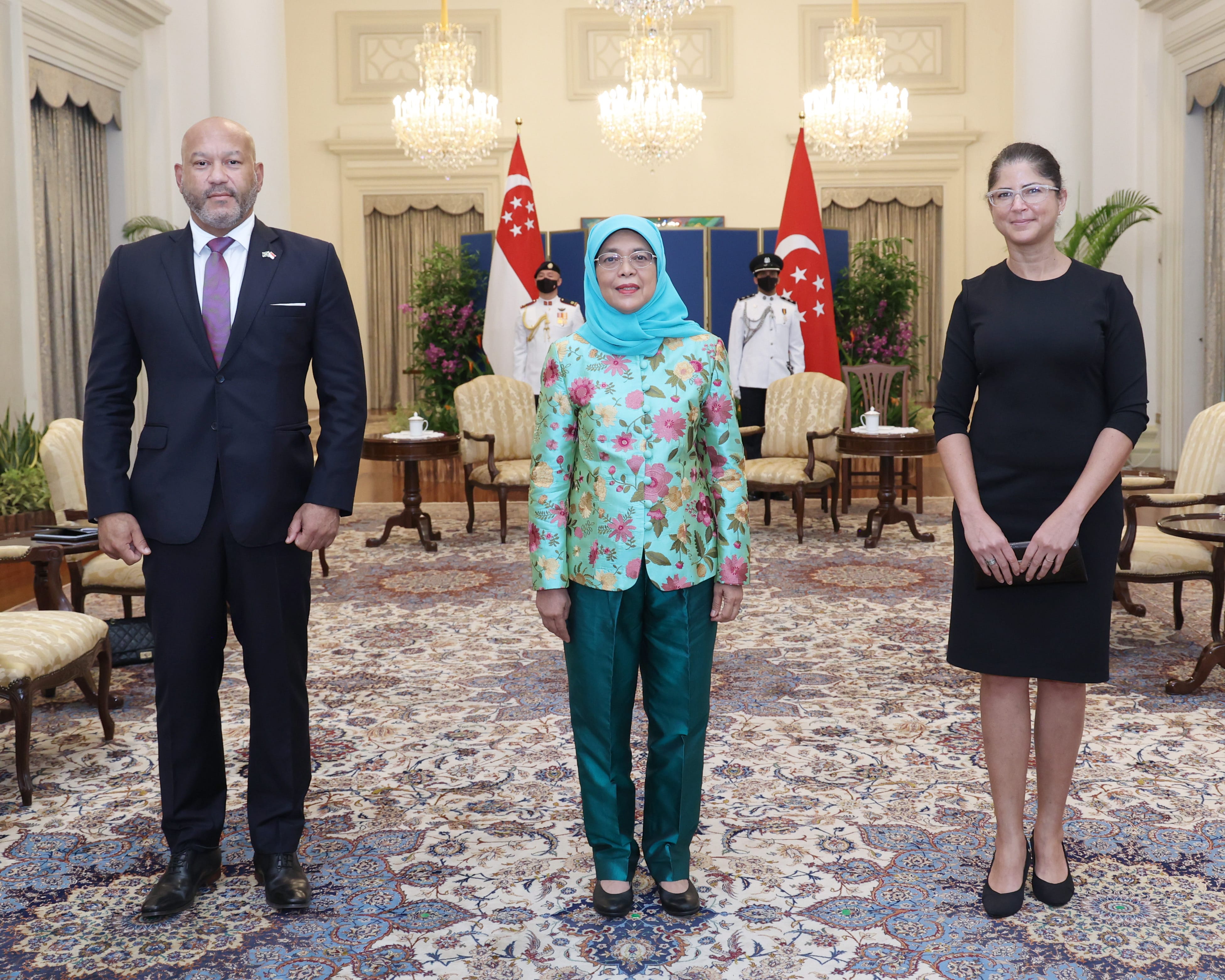 Halimah Yacob stands between two people, Singapore flags behind, ornate room.