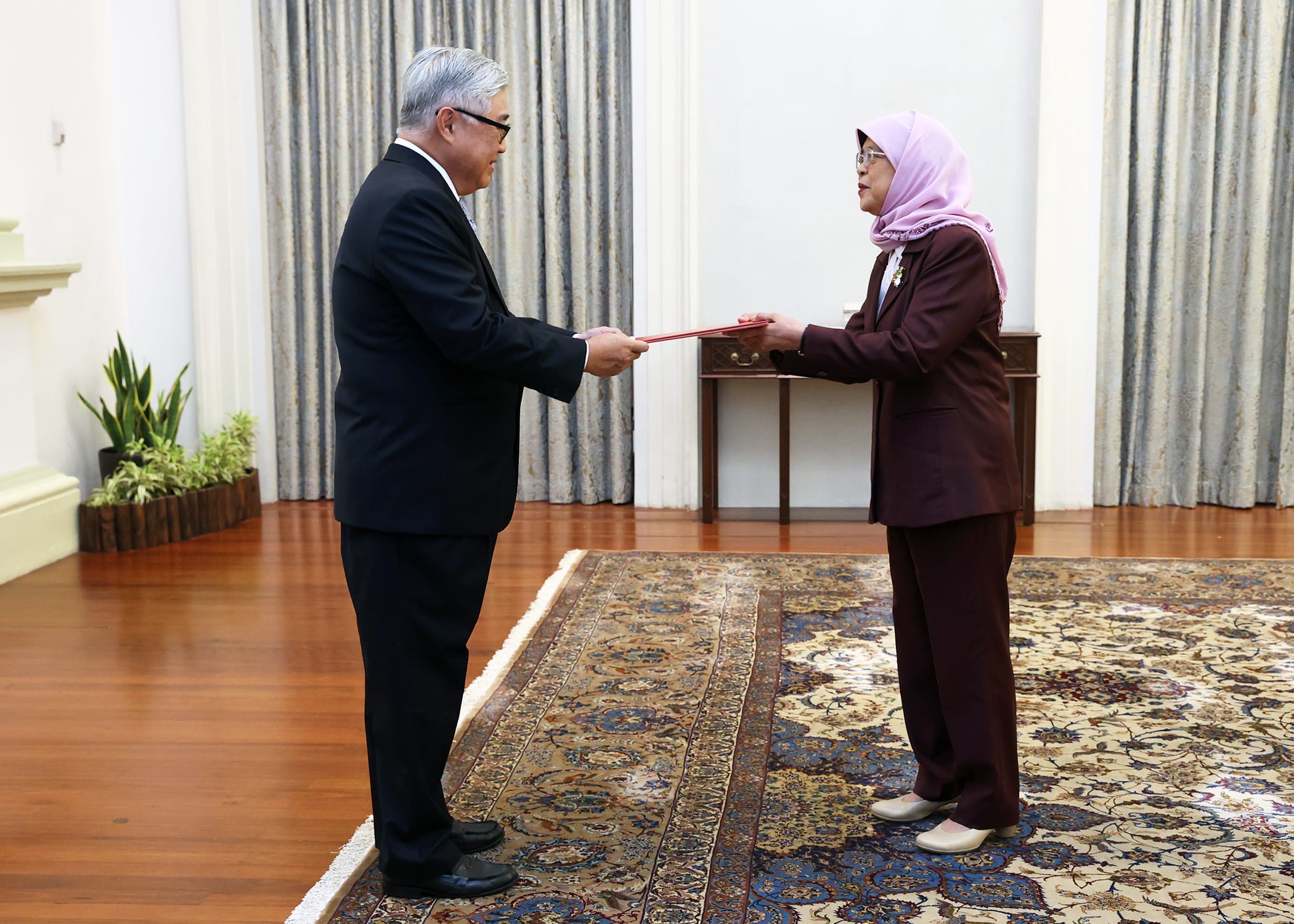 Two people exchanging a red document, ornate rug on floor, formal setting.