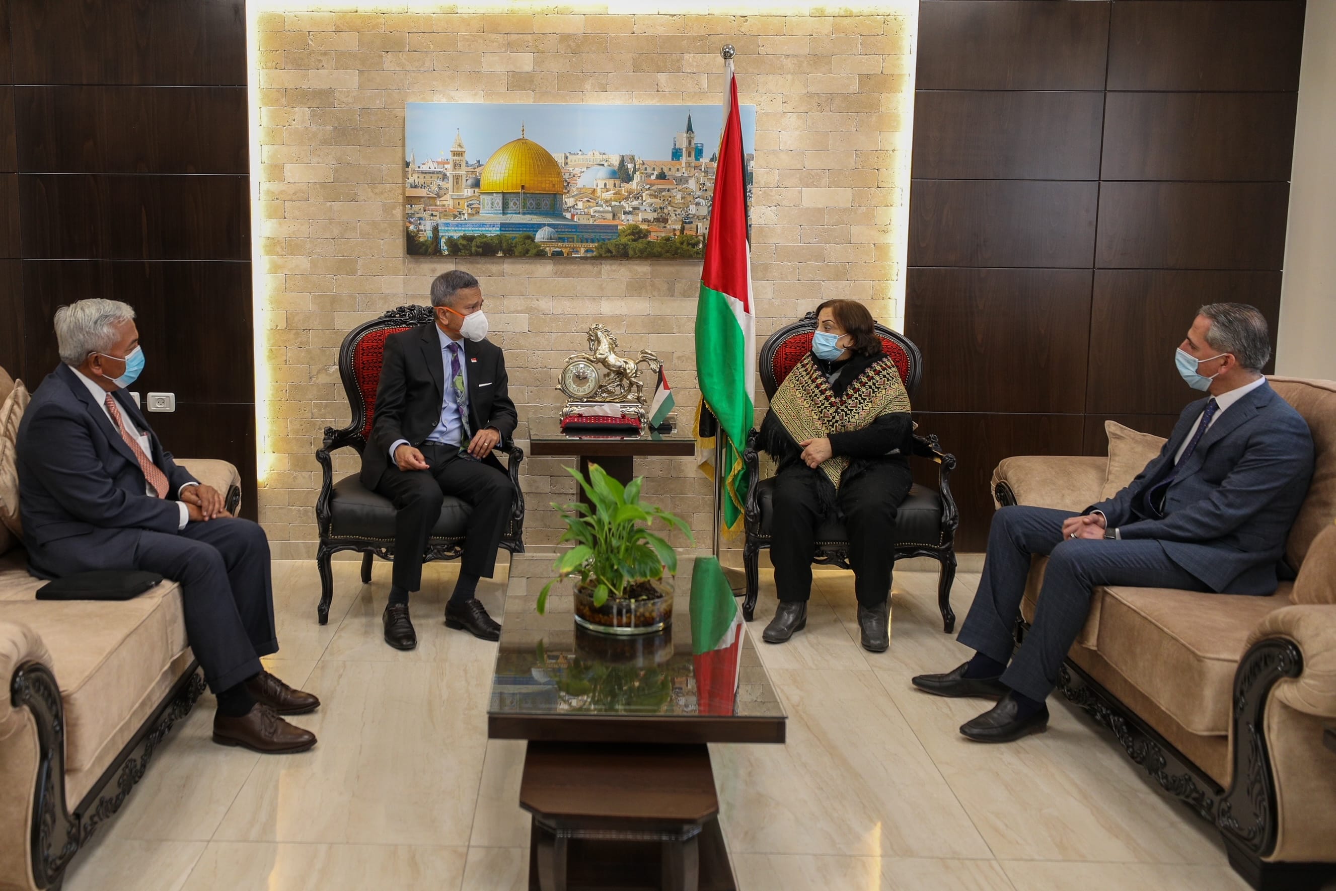 Four people in suits wearing masks sit in a room with the Palestinian flag and a Dome of the Rock image.