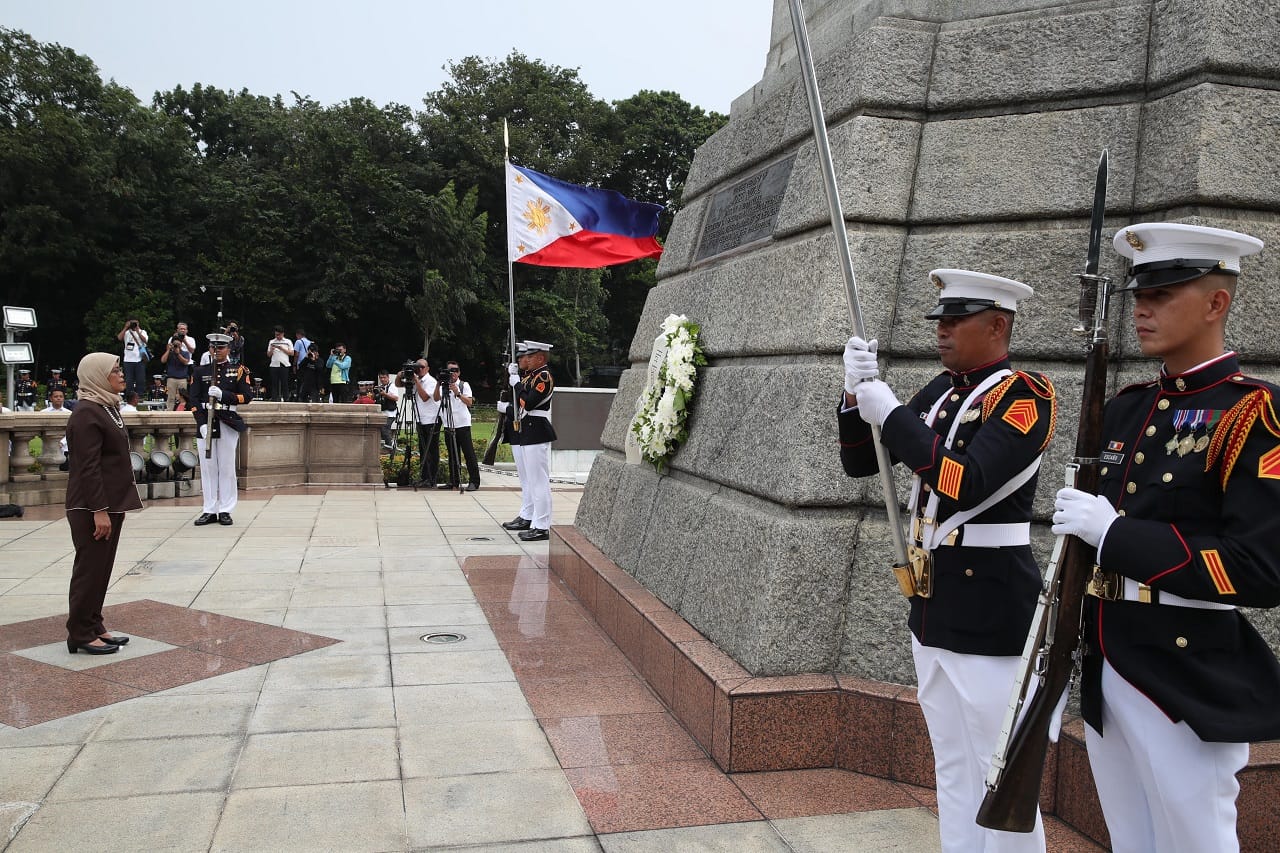 Military honor guard at monument with Philippines flag, woman in suit, photographers in background.