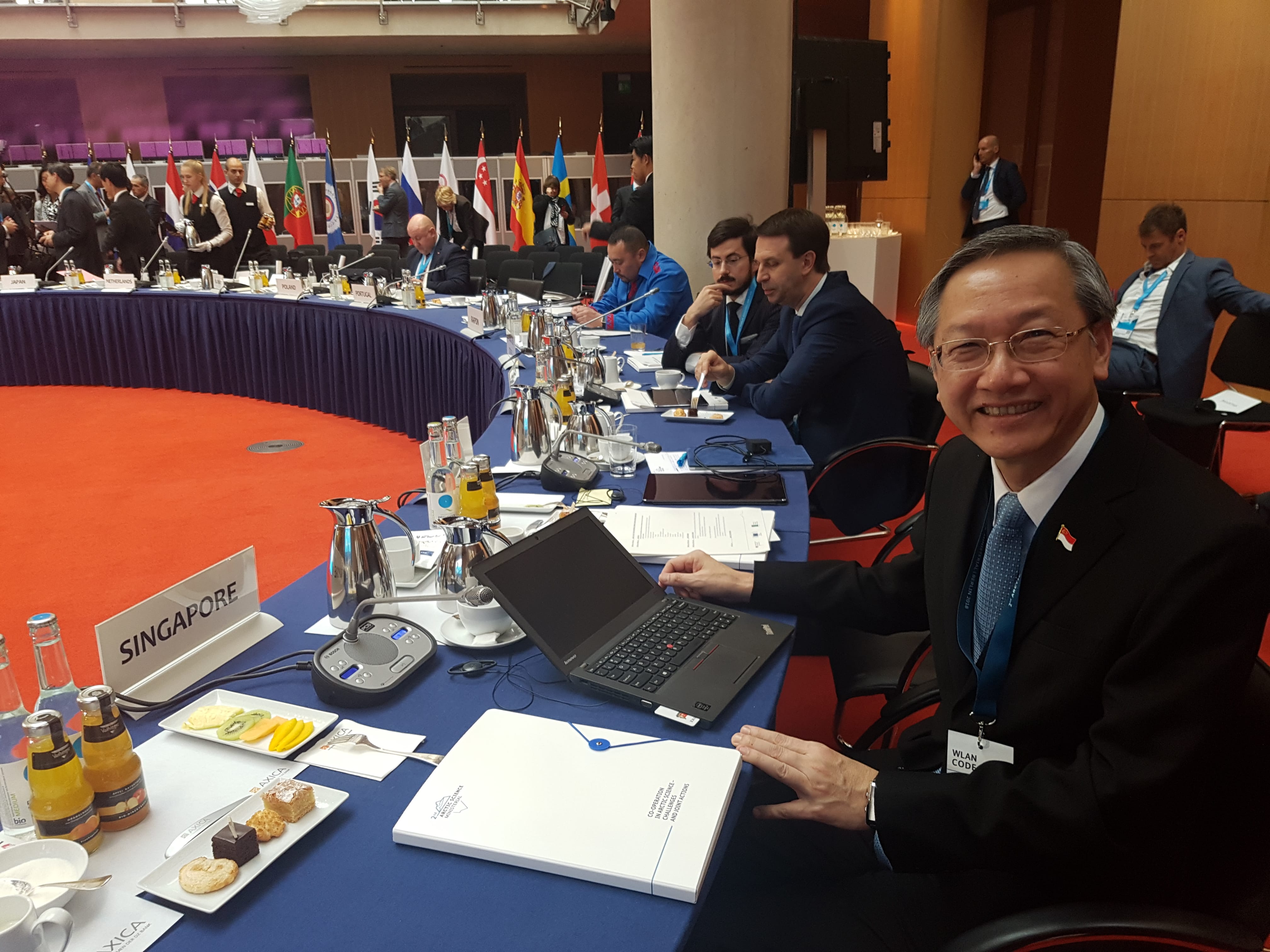 Person at "Singapore" table with laptop in meeting room; flags line the back.