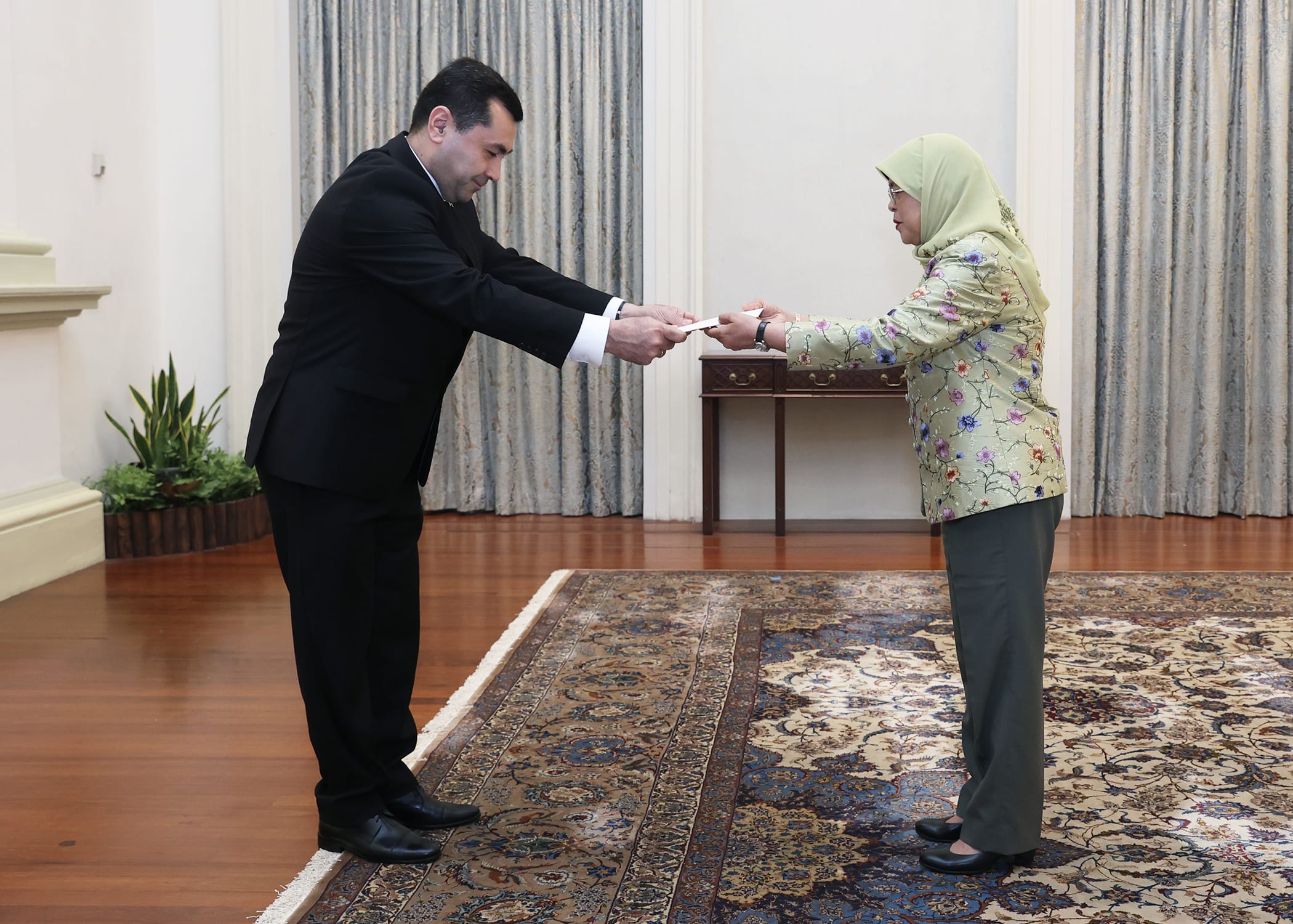 Two people exchanging a document, one in a suit, the other in floral jacket and hijab.