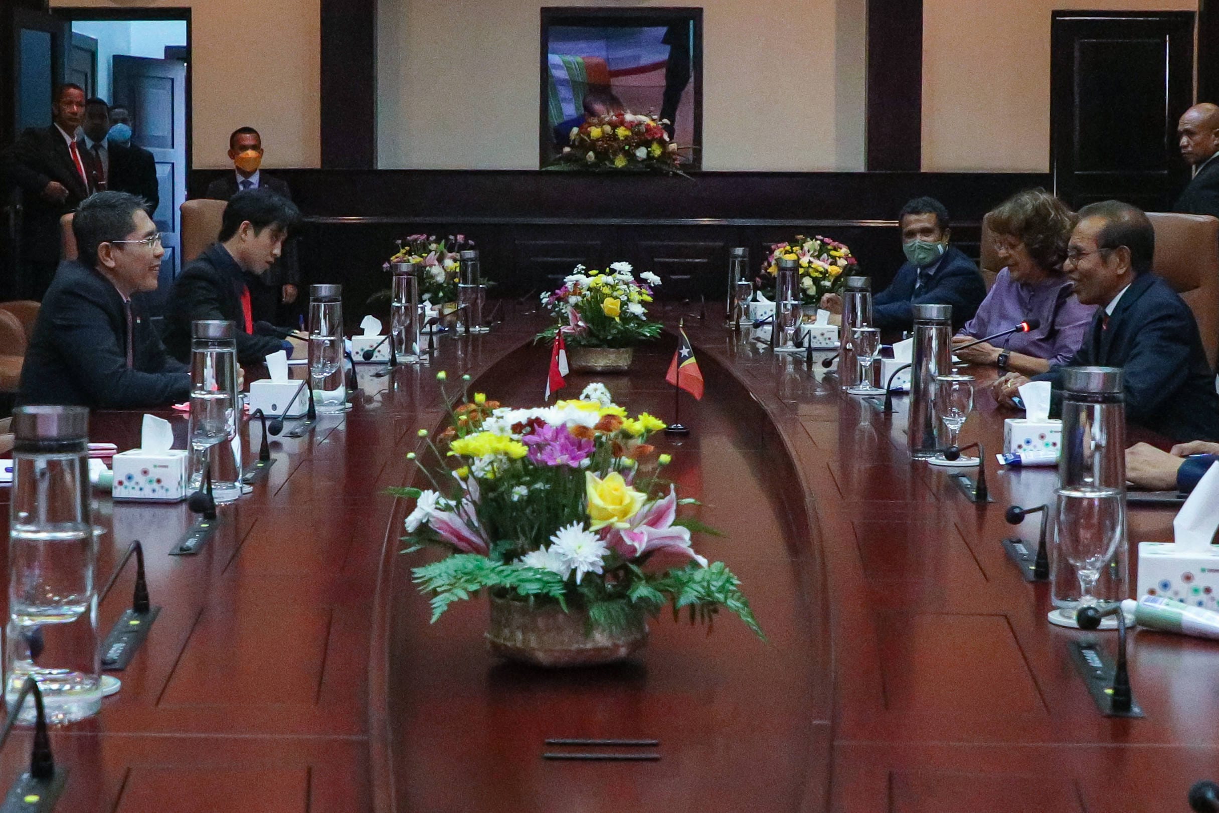 People seated at a long conference table with microphones, water, tissues, and flower arrangements.