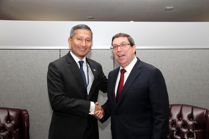 Two men in suits shake hands in front of a gray wall.