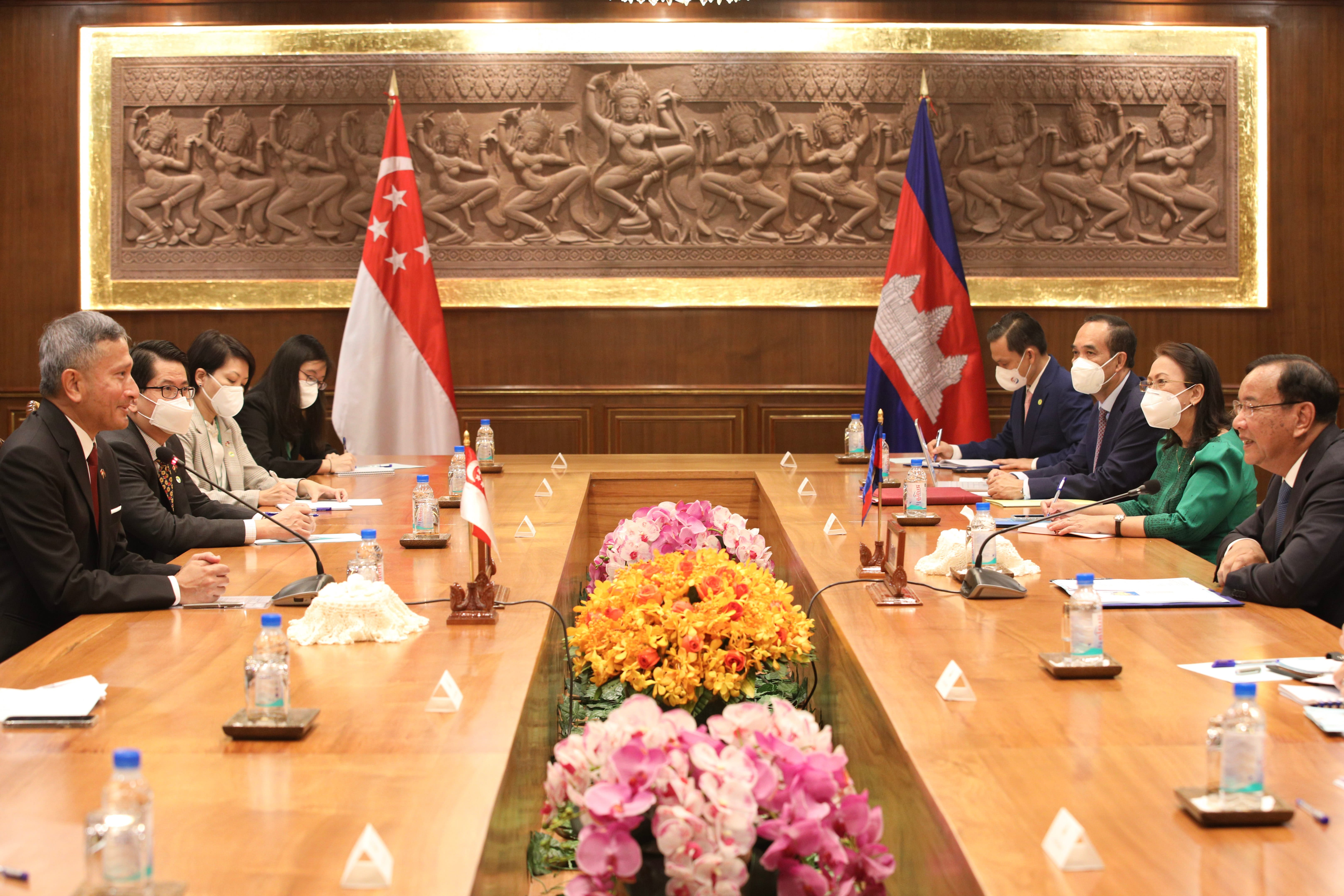 Singapore and Cambodia flags at meeting table with masked officials, carvings on the wall.