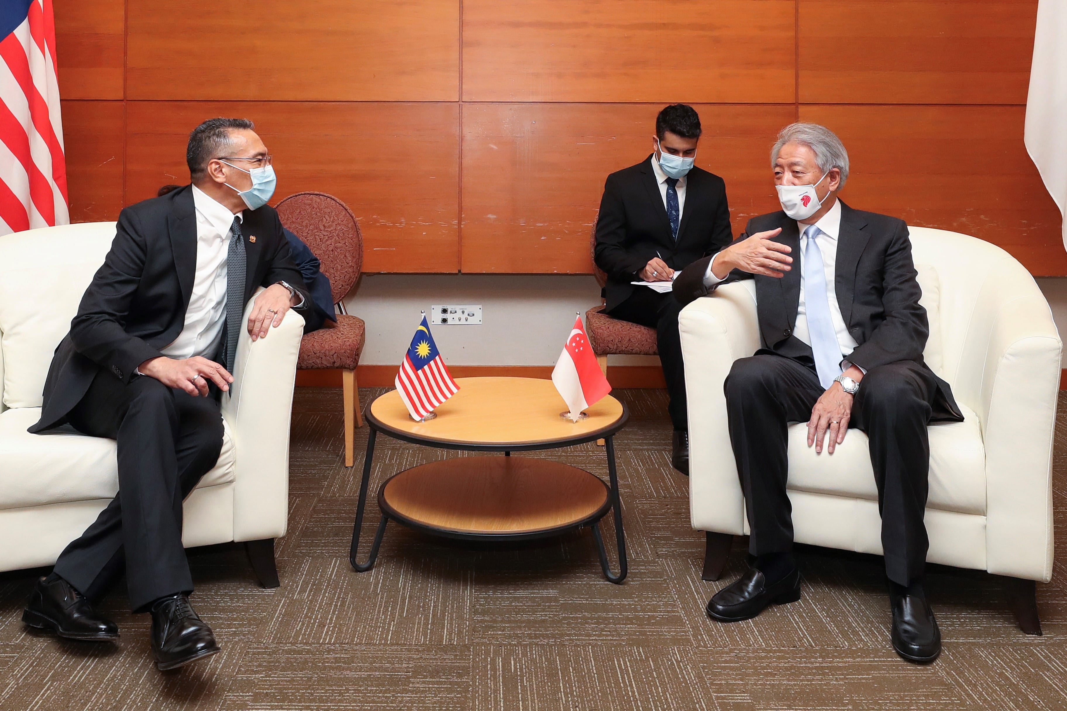Two suited men with masks sit in armchairs; flags of Malaysia and Singapore on table.