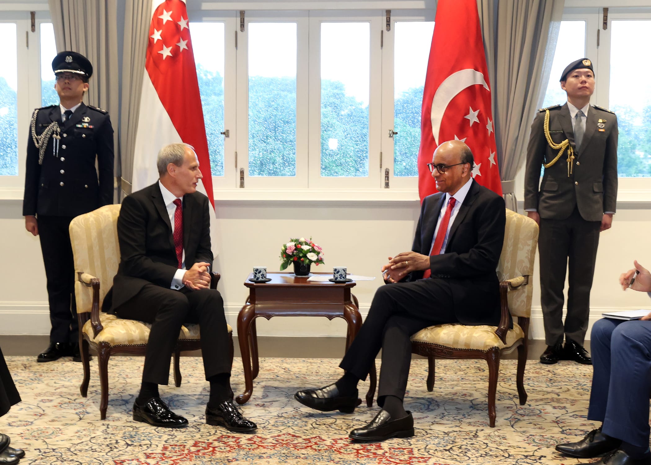 Two men in suits sit, facing each other with Singapore flags and guards standing behind them.