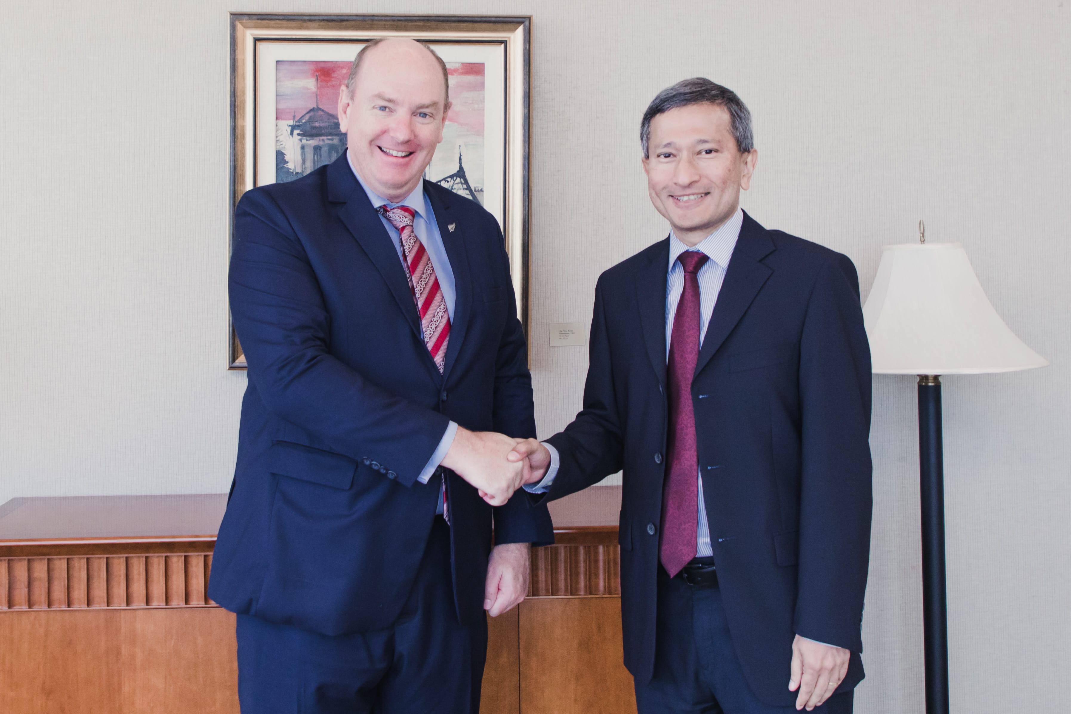 Two men in suits shaking hands, with artwork and lamp visible.