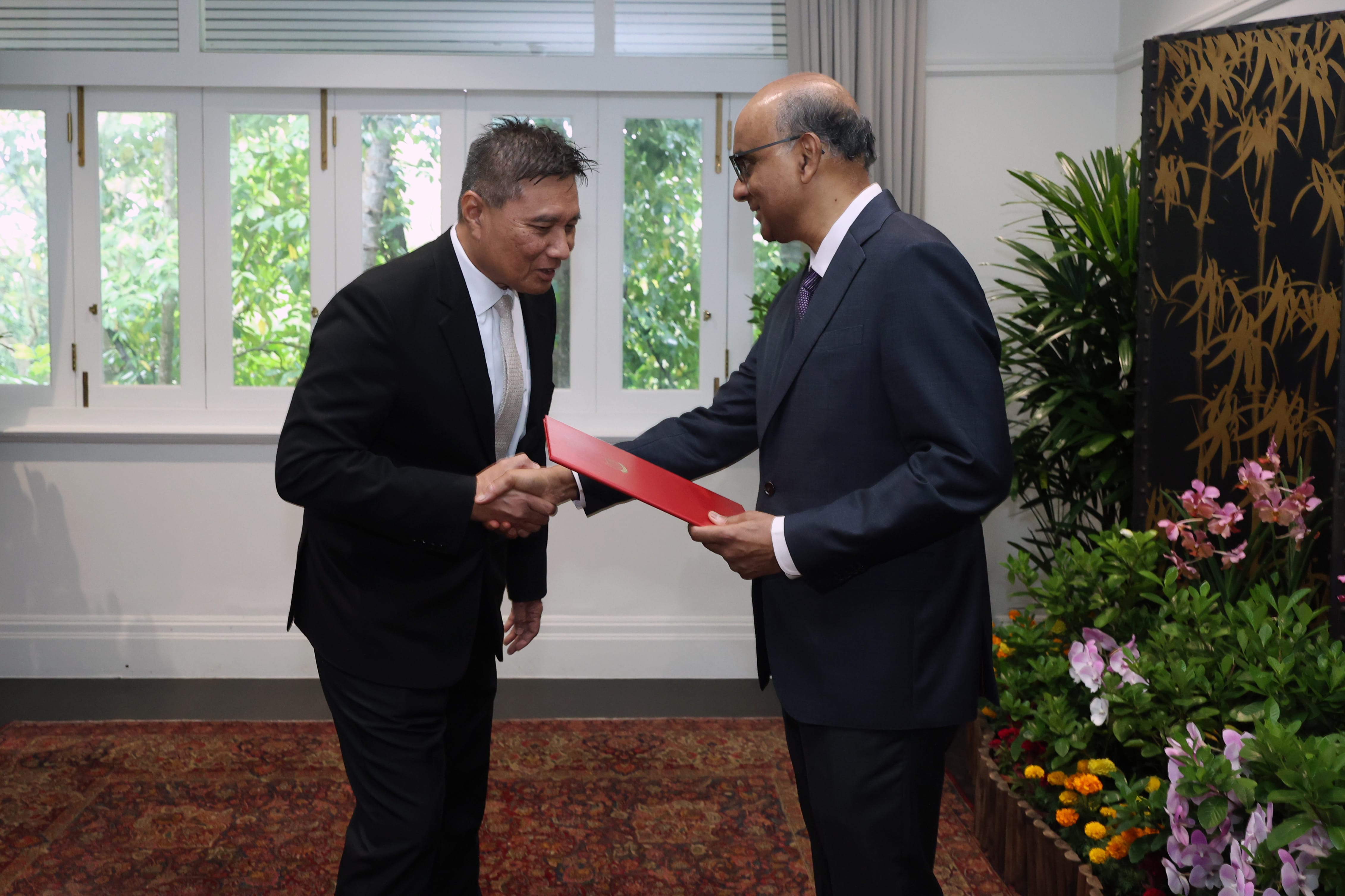 Two men in suits shaking hands, one holding a red folder. Window and flower arrangement behind.