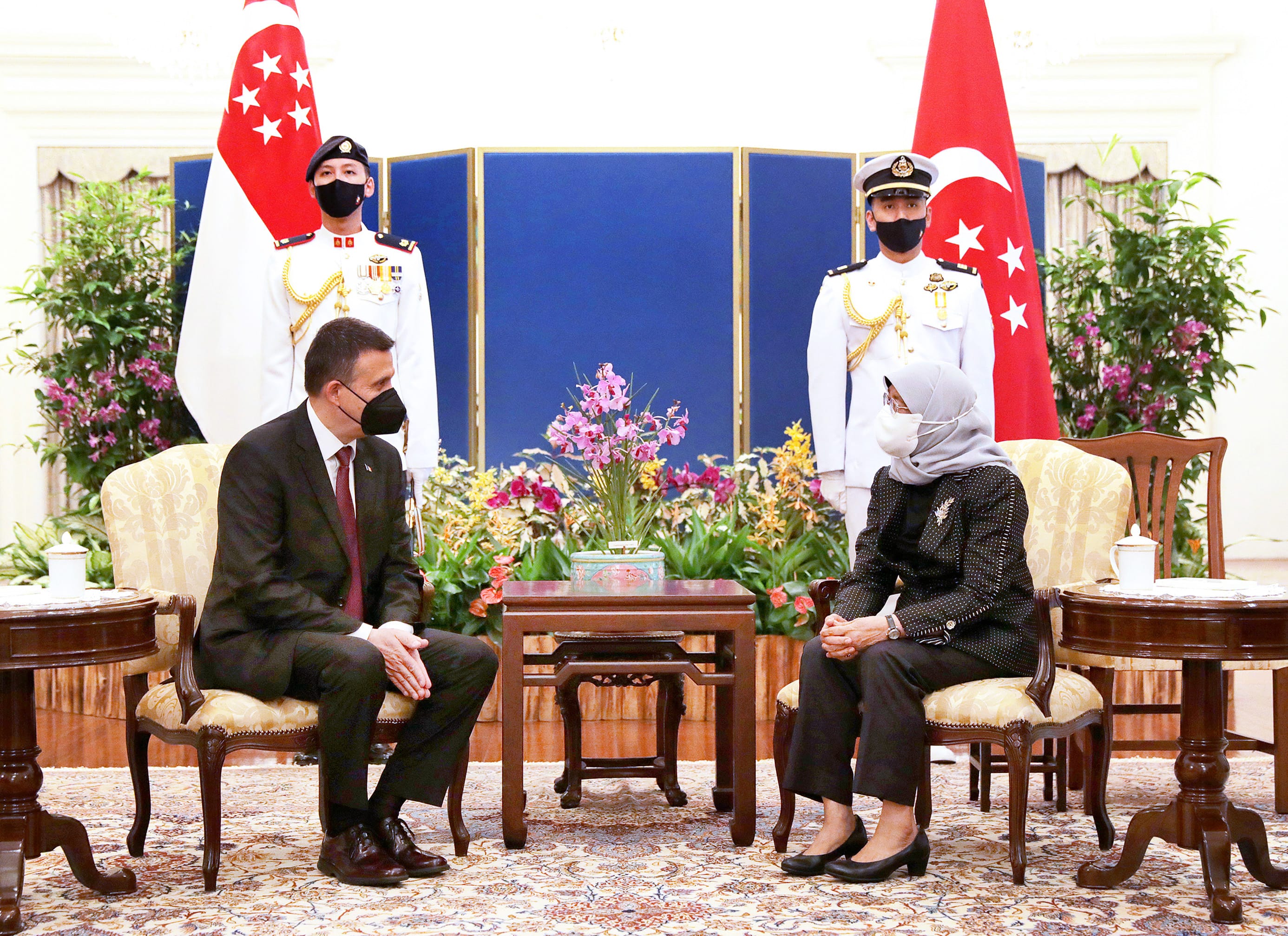 Man and woman with face masks seated; Singapore flags and guards in the background.