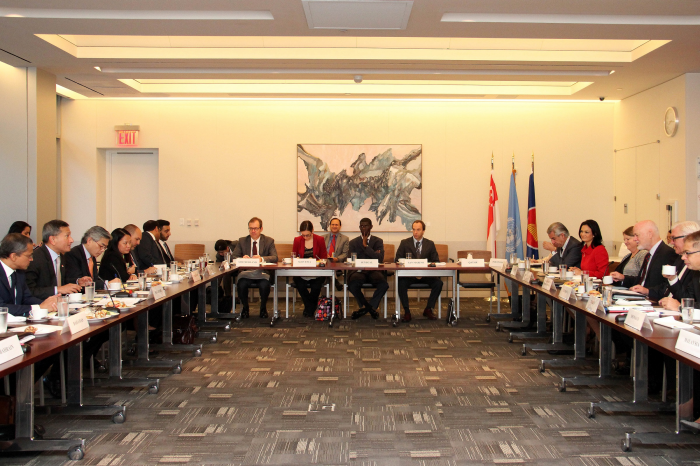 Conference room with people seated at two long tables, flags in the background.