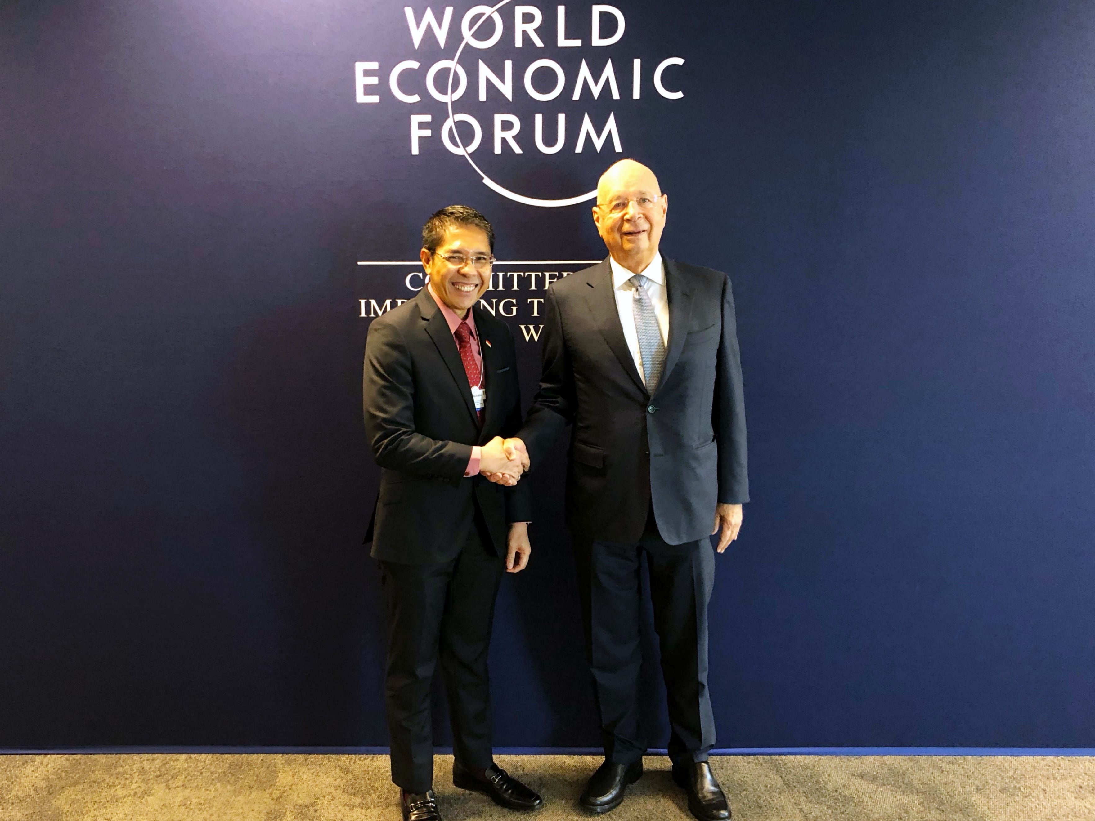 Two men hold a gift-wrapped box in a room with framed portraits and Kuwaiti flag.