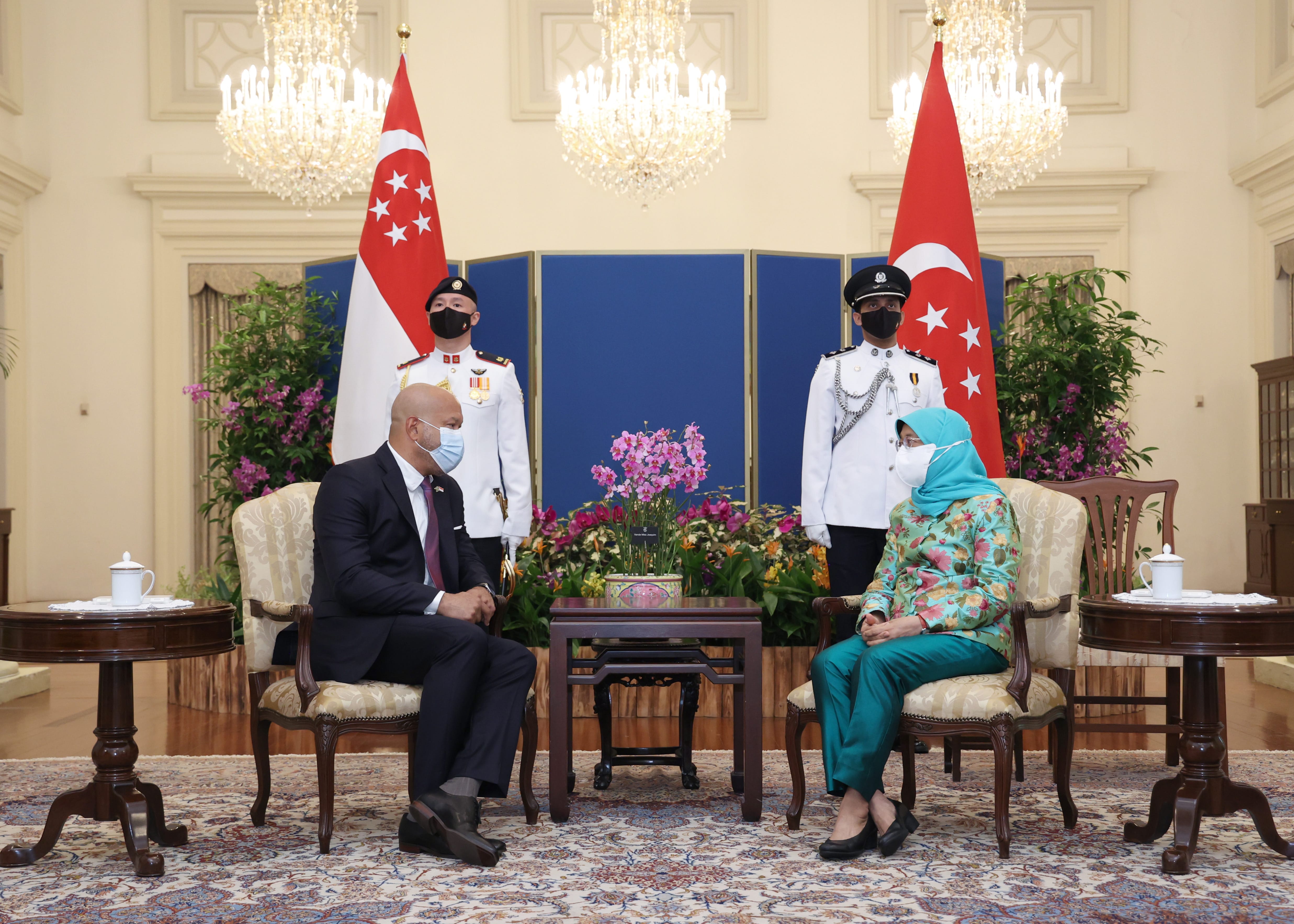 Singapore President Halimah Yacob and a man sit in chairs, masked, with flags and honor guard.
