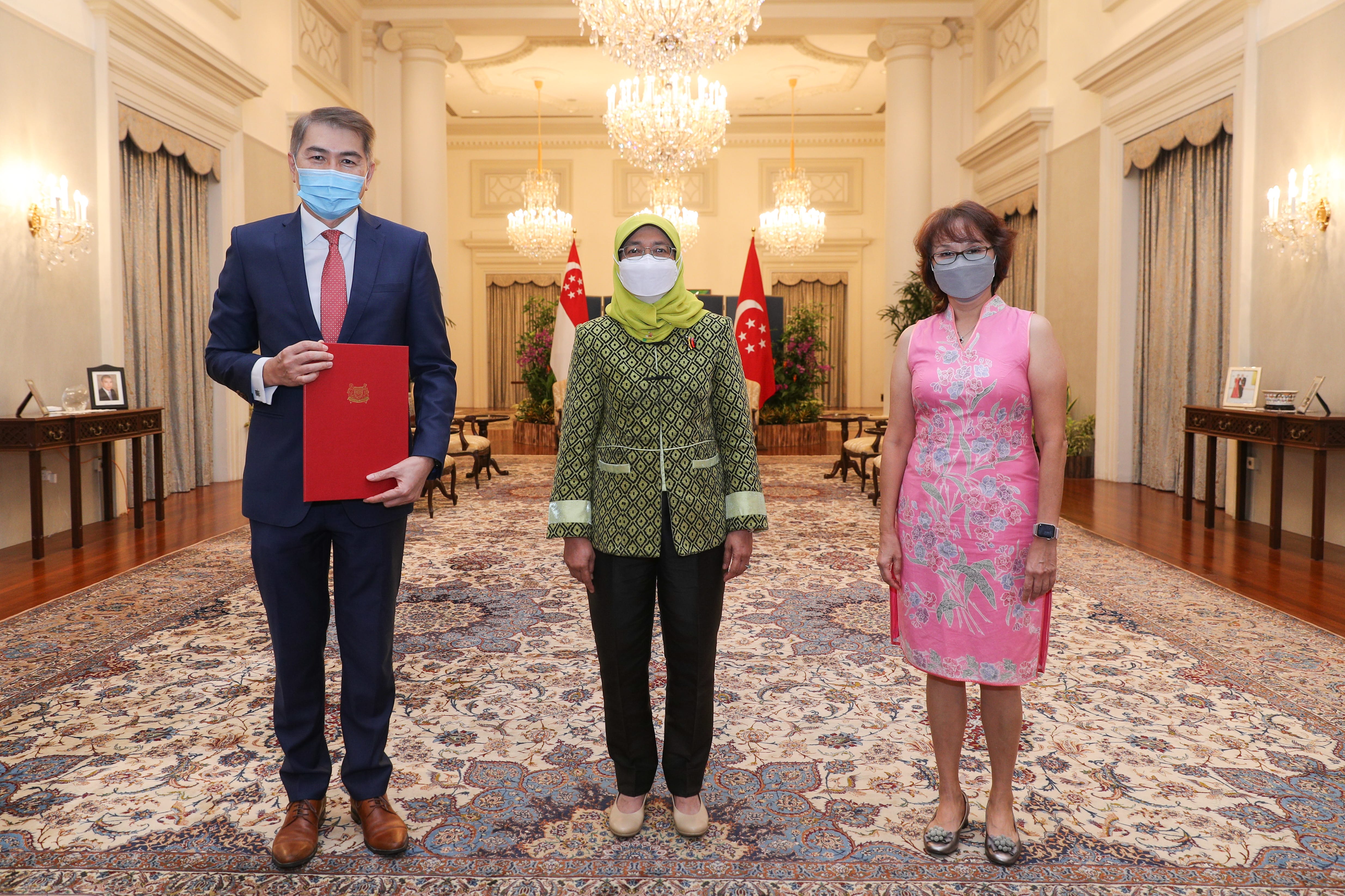 Three masked people pose indoors before a Singapore flag, chandelier, and ornate carpet.