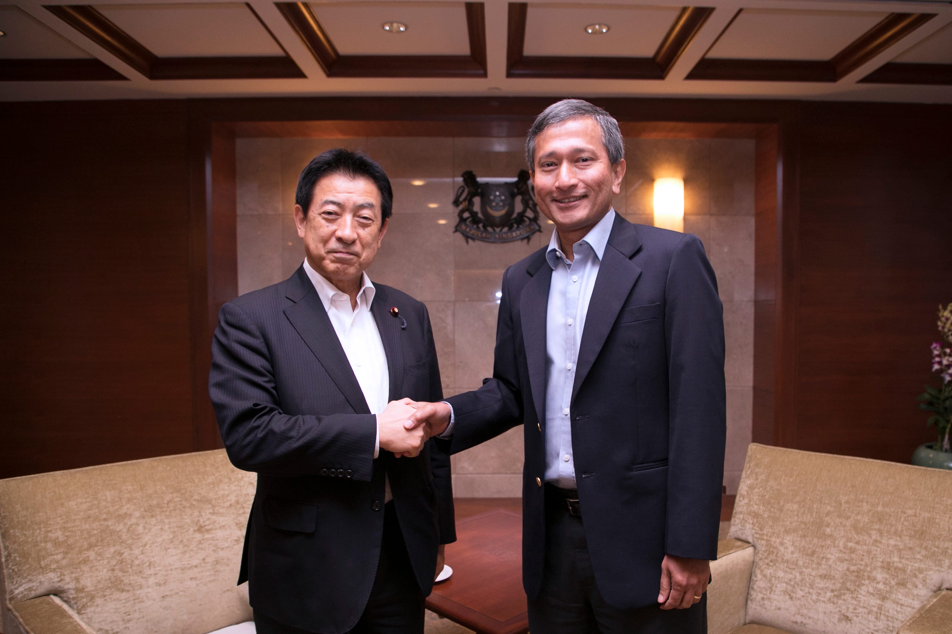 Two men in suits shake hands in front of a wall with a coat of arms.