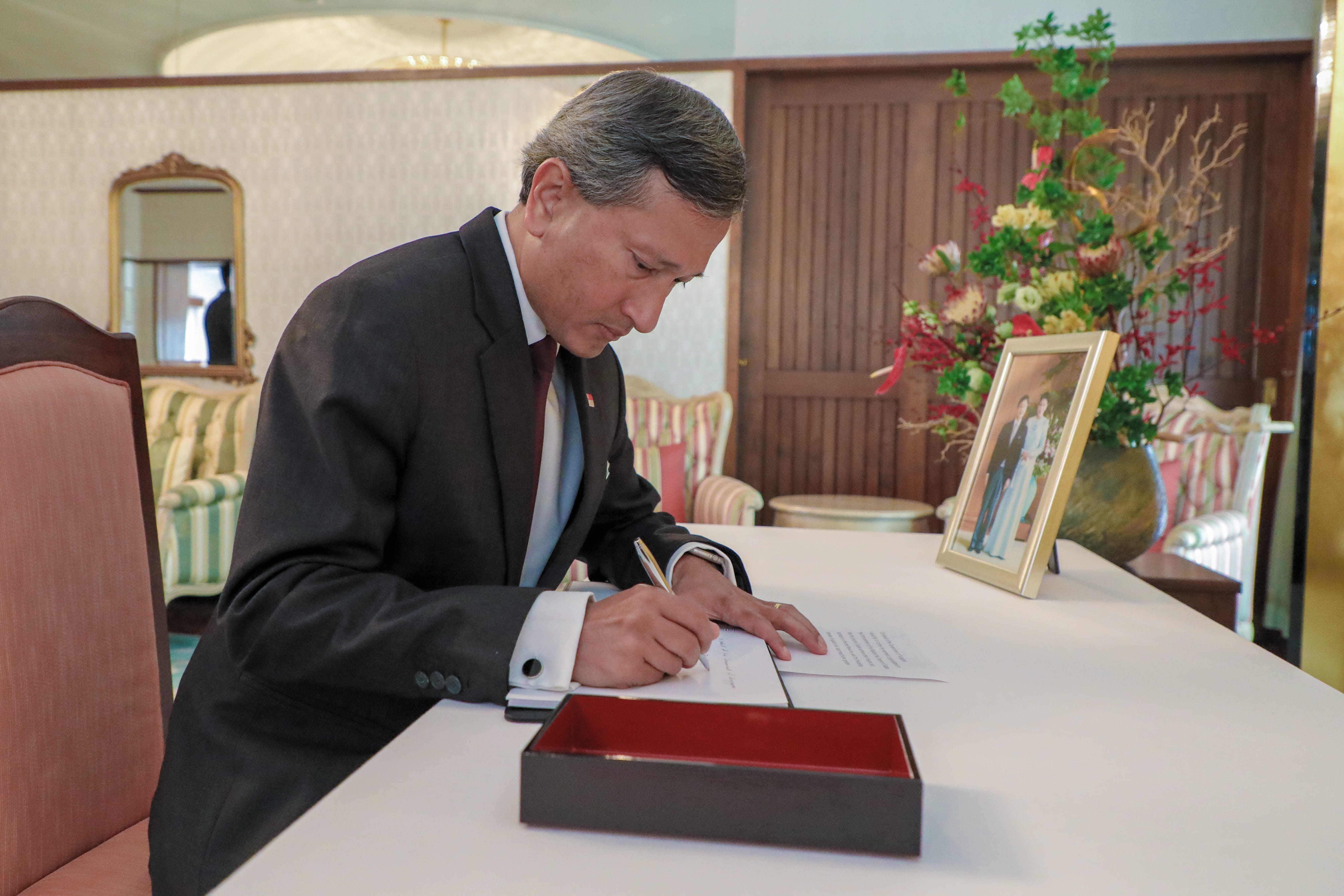 Man in suit signing document at table with box, flowers, and framed photo.