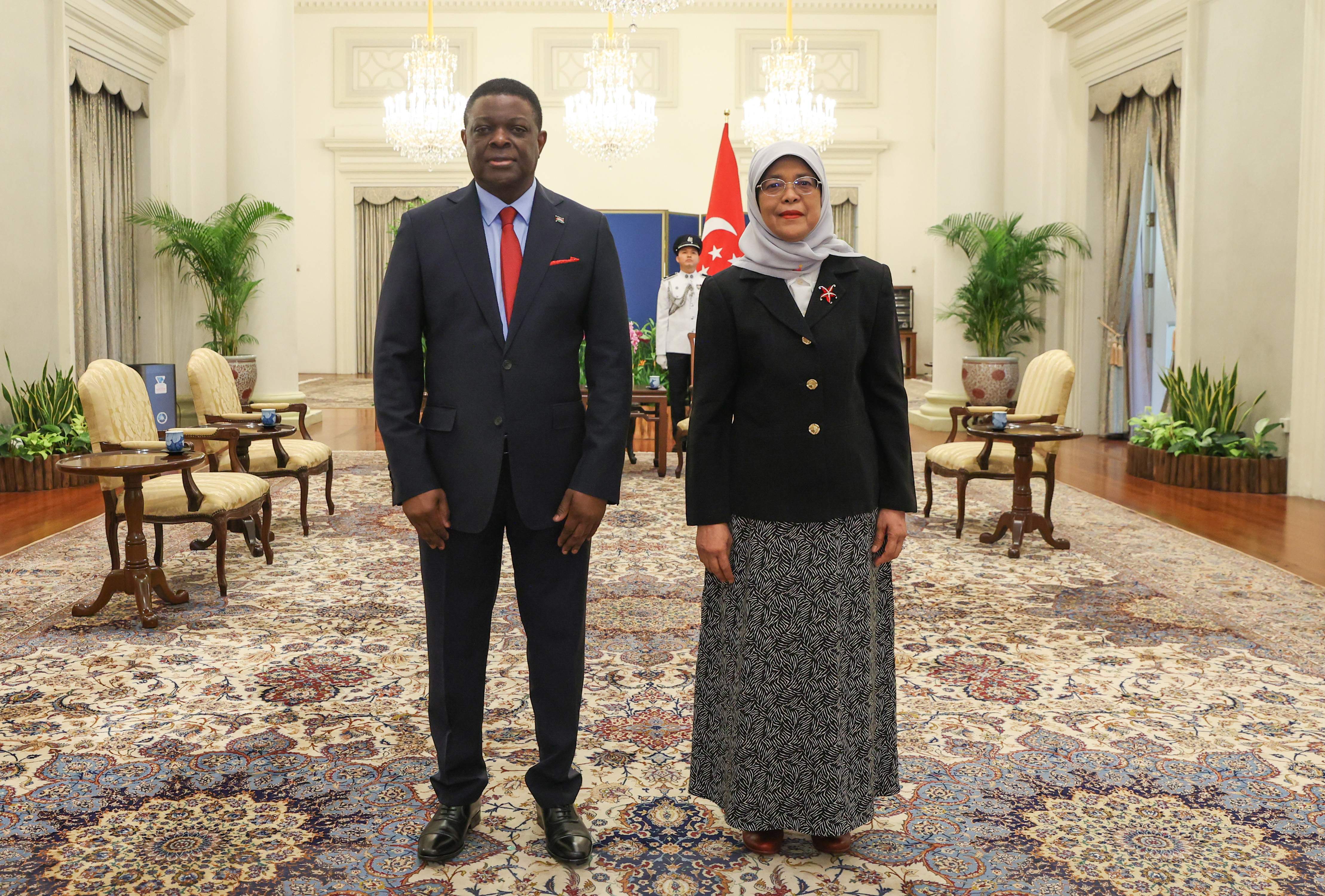 Man in suit with red tie and Halimah Yacob, President of Singapore, indoors, guard behind them.