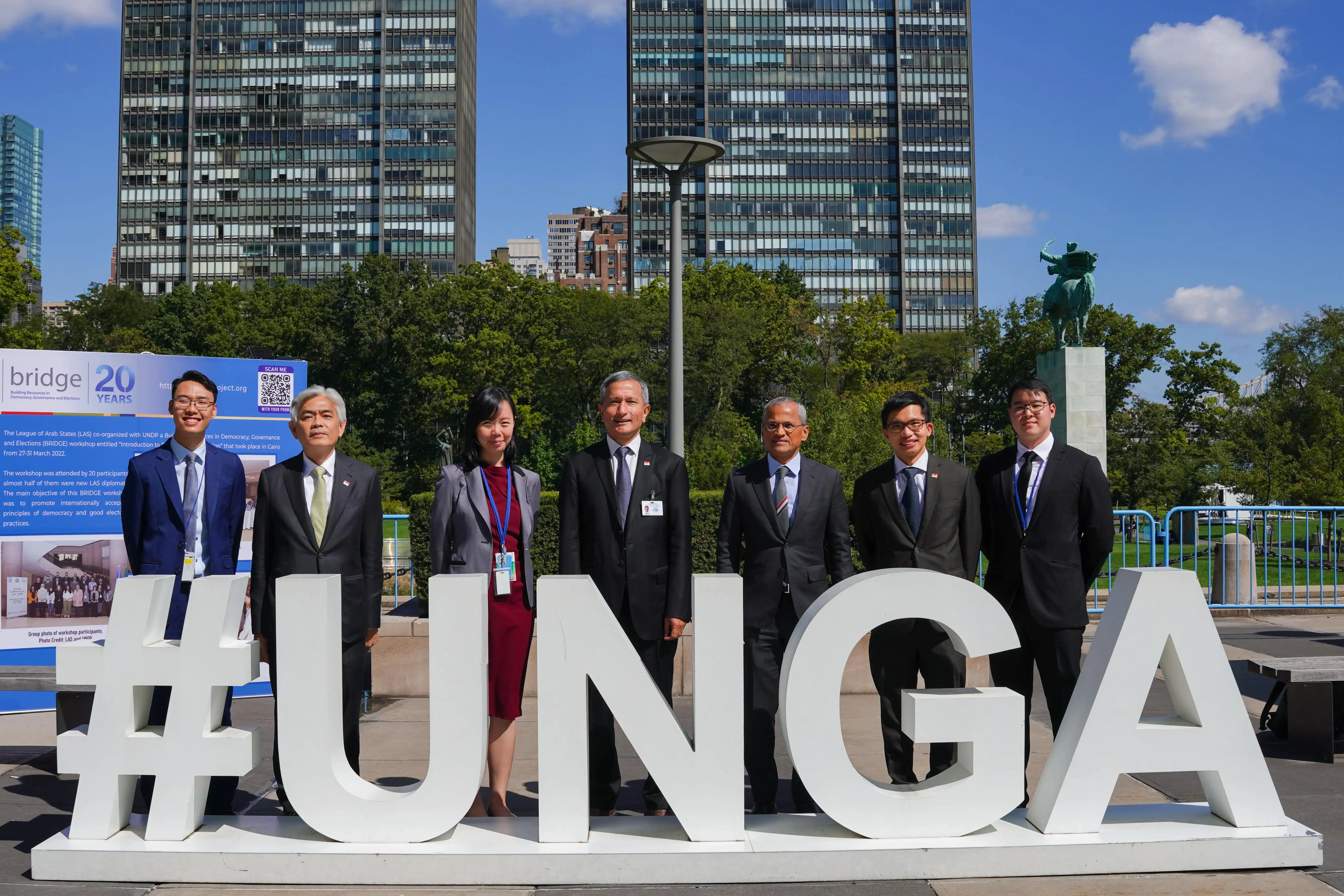 Group of seven people in formal wear posing in front of large "#UNGA" letters at an open ground.