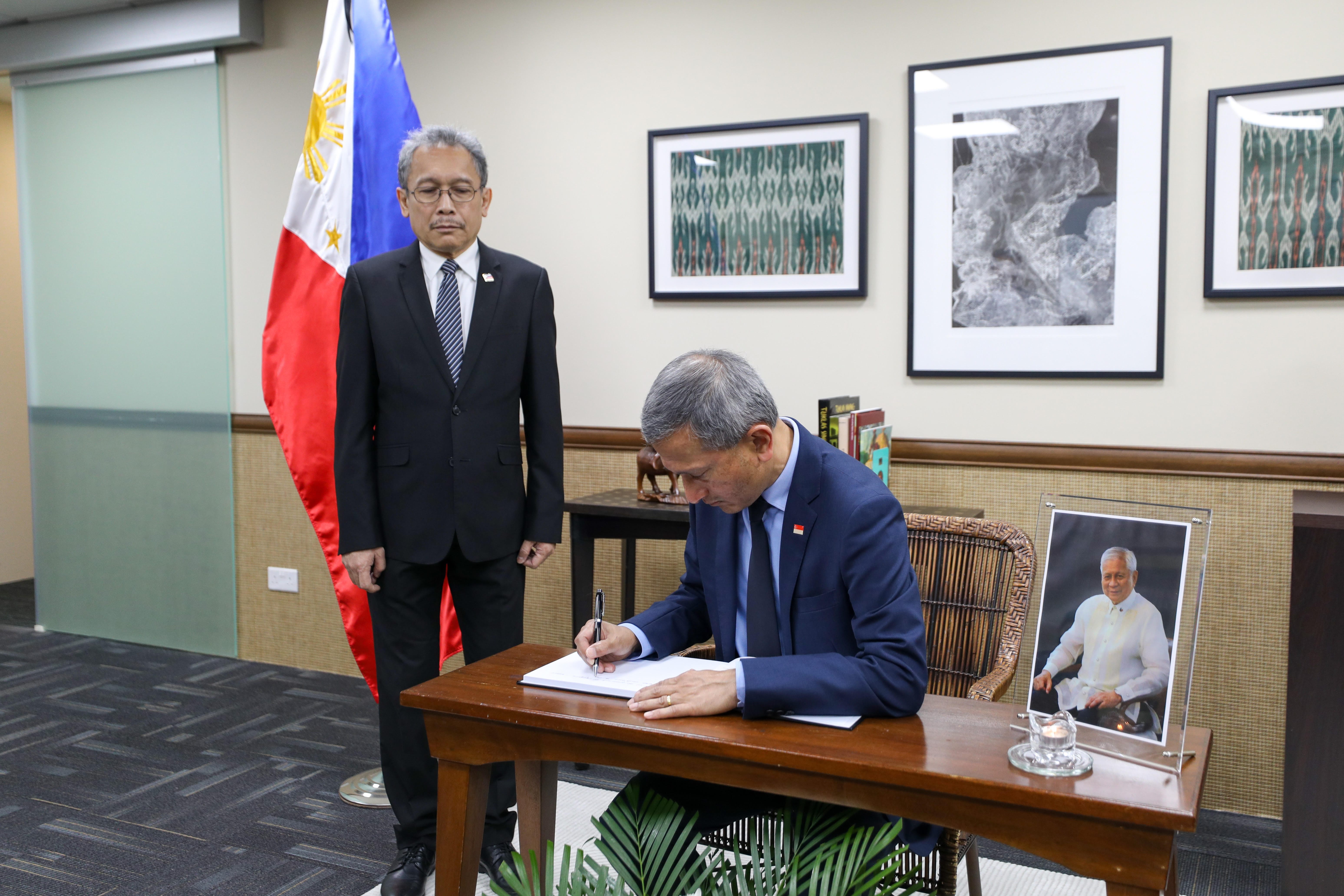Two men; one in suit signing document, other standing by Philippines flag. Framed photo visible.
