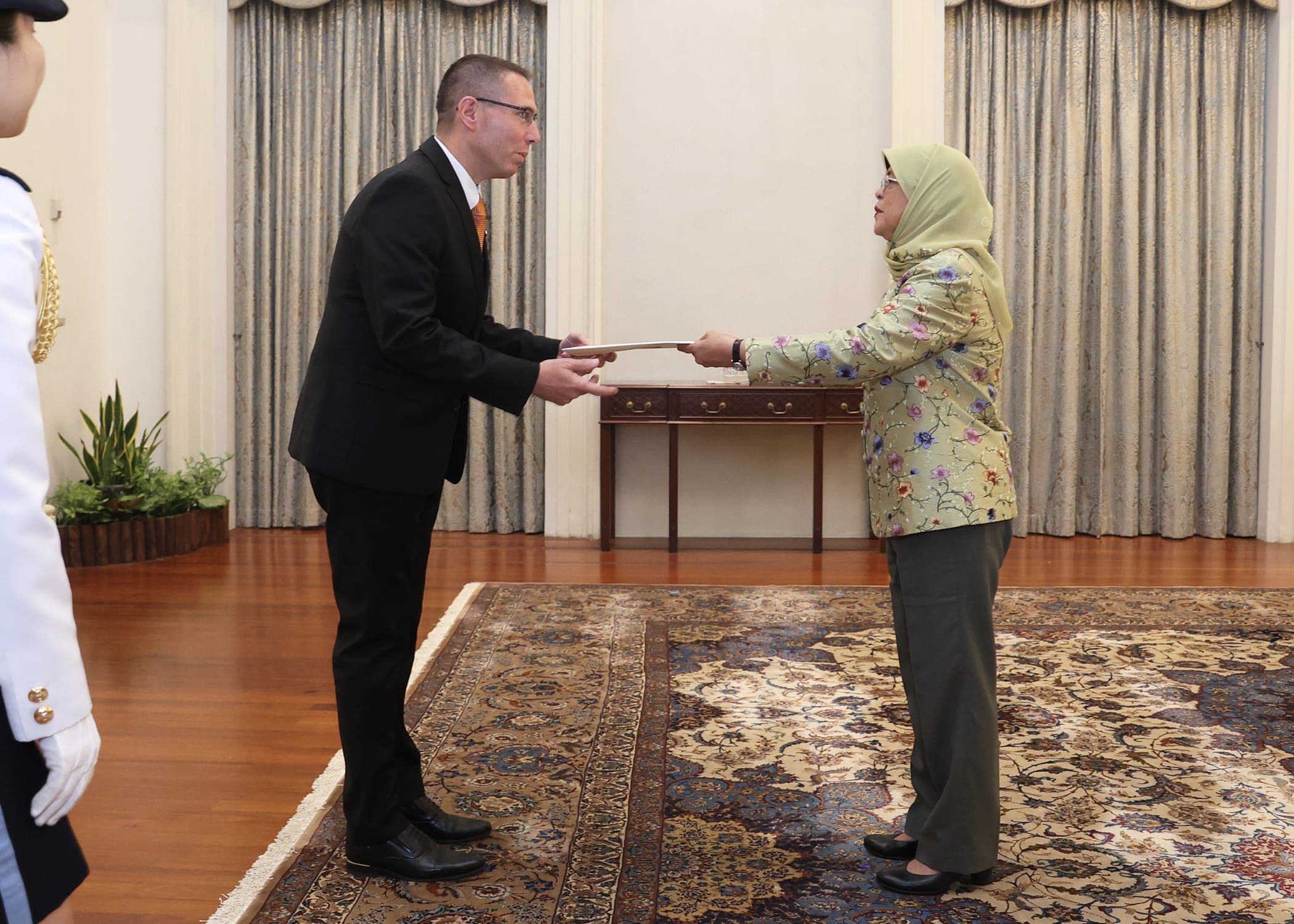 Man in suit presents document to woman in headscarf; patterned rug, formal room.
