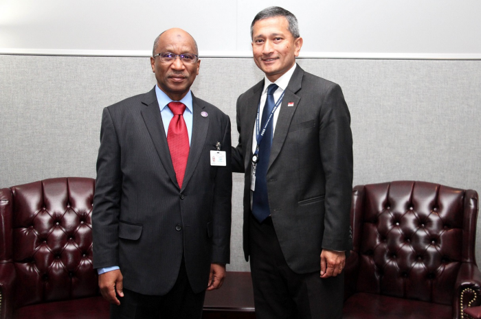 Two men in suits pose near burgundy armchairs against a gray wall.