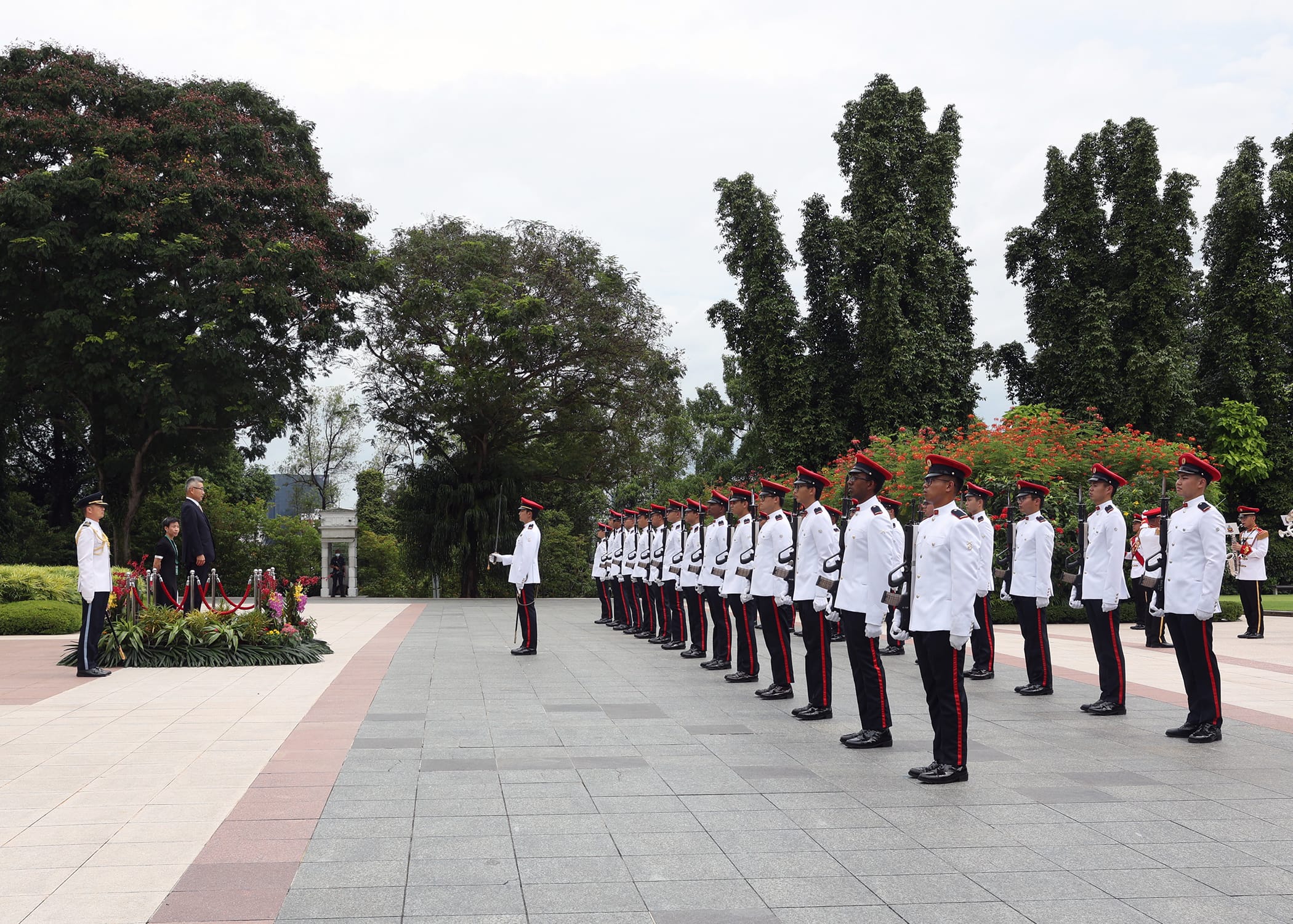 Honor guard in dress uniforms stands at attention facing dignitaries by floral arrangement.