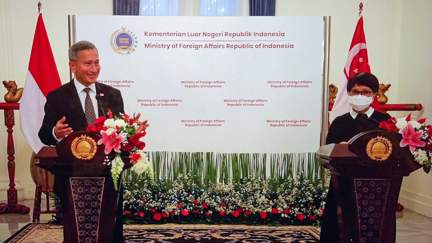Woman in pink, wearing a mask, signs a document on a red-topped desk near a window.